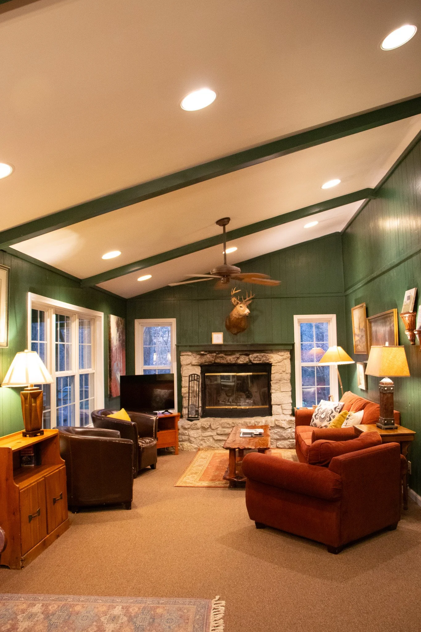 Cozy living room with green paneled walls, stone fireplace, mounted deer head, ceiling fan, leather and fabric sofas, side tables with lamps, flat-screen TV, and decorative art.