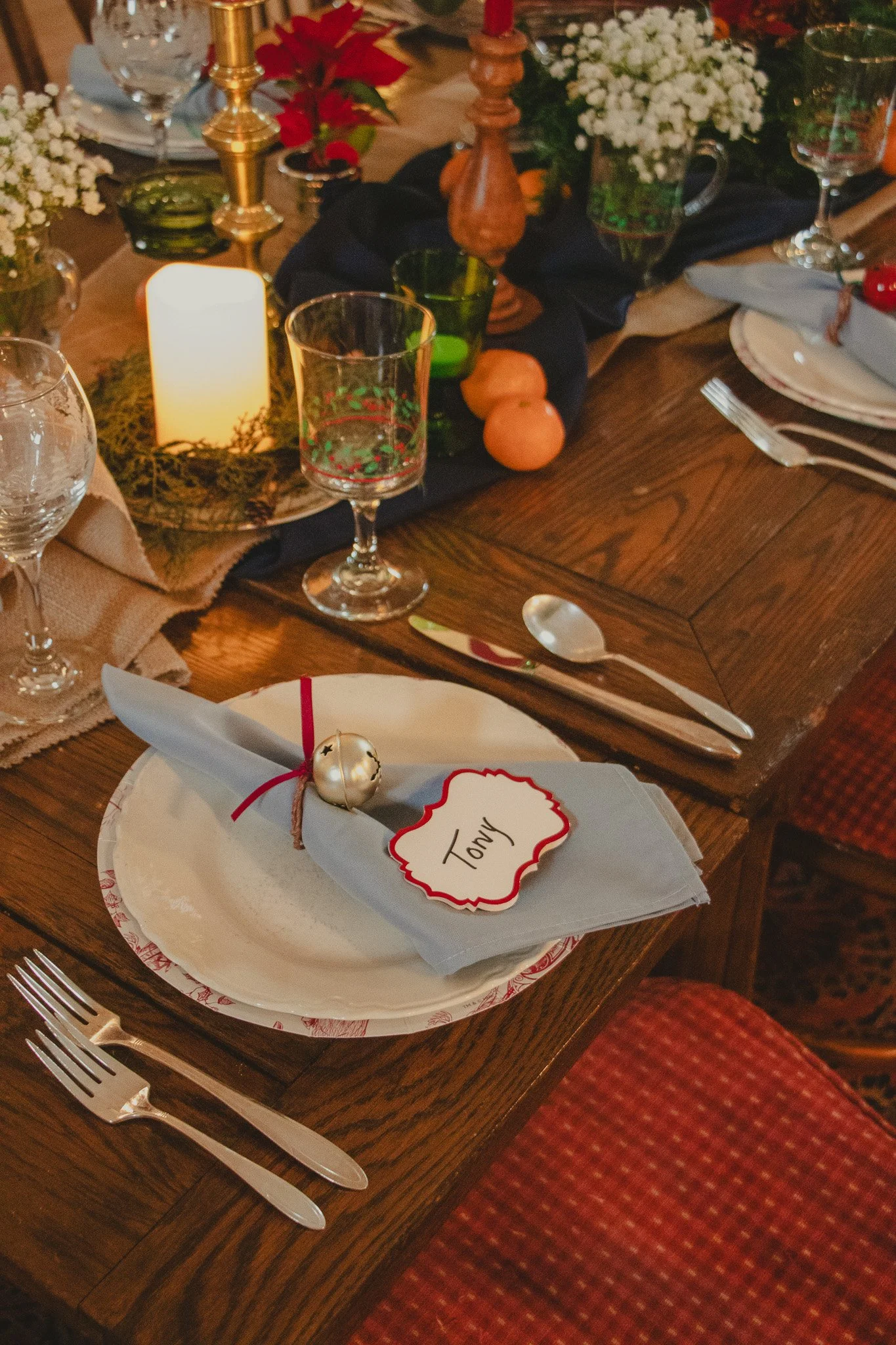 A holiday table setting with a white plate, a folded napkin decorated with a small jingle bell and a name tag labeled 'Tony', surrounded by silverware, glasses, candles, and festive decorations including flowers and fruits.
