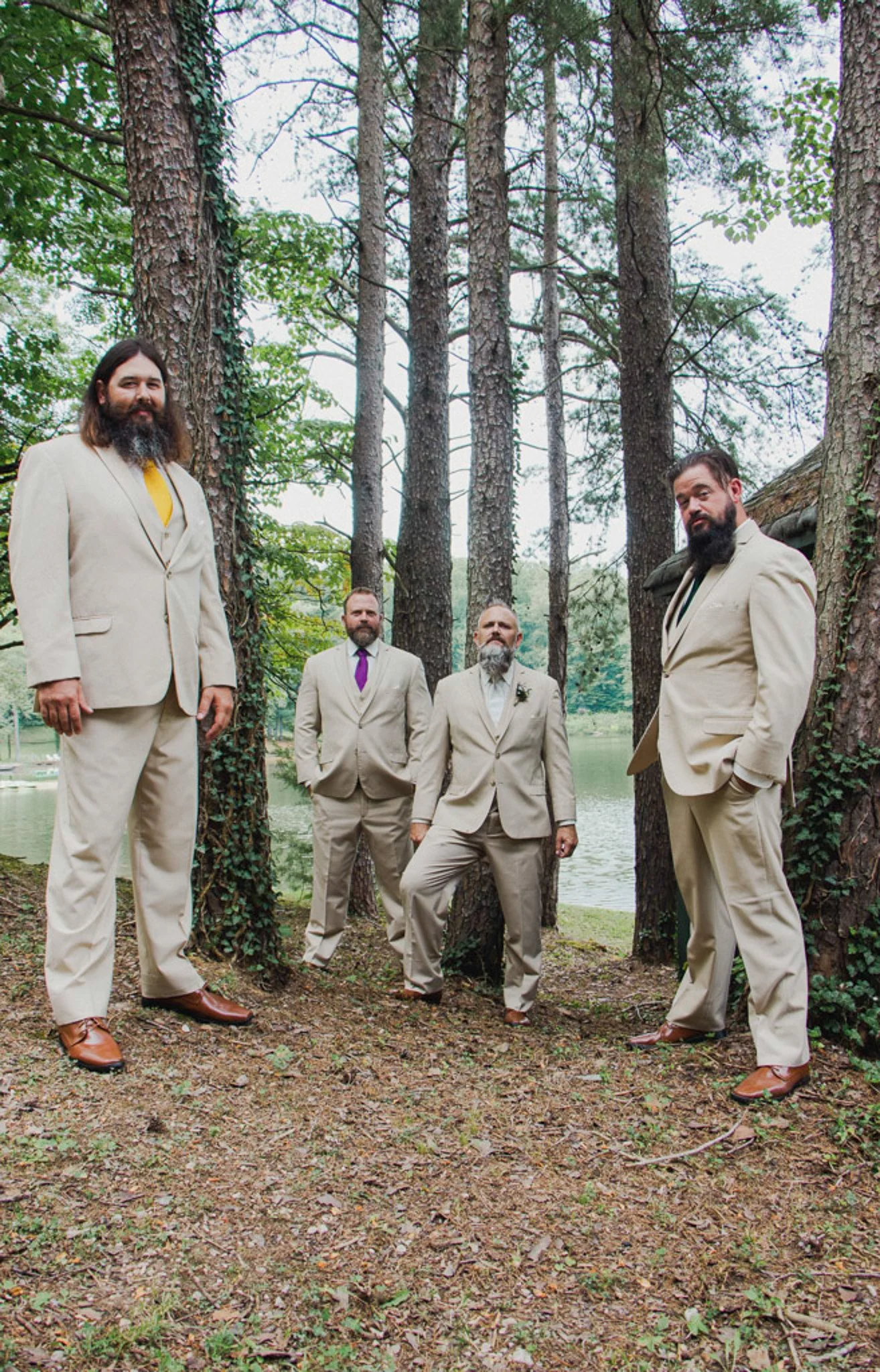Four men dressed in beige suits pose outdoors beside trees near a body of water, with tall pine trees and an overcast sky in the background.