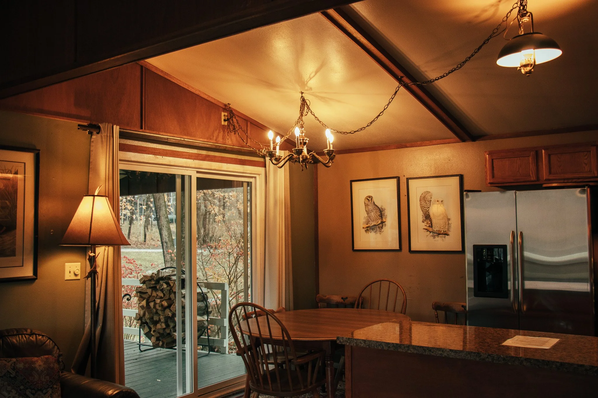 A cozy kitchen and dining area with wood accents, a sliding glass door leading to a deck with firewood, framed owl artwork on the wall, a metallic refrigerator, and a chandelier hanging from chains.