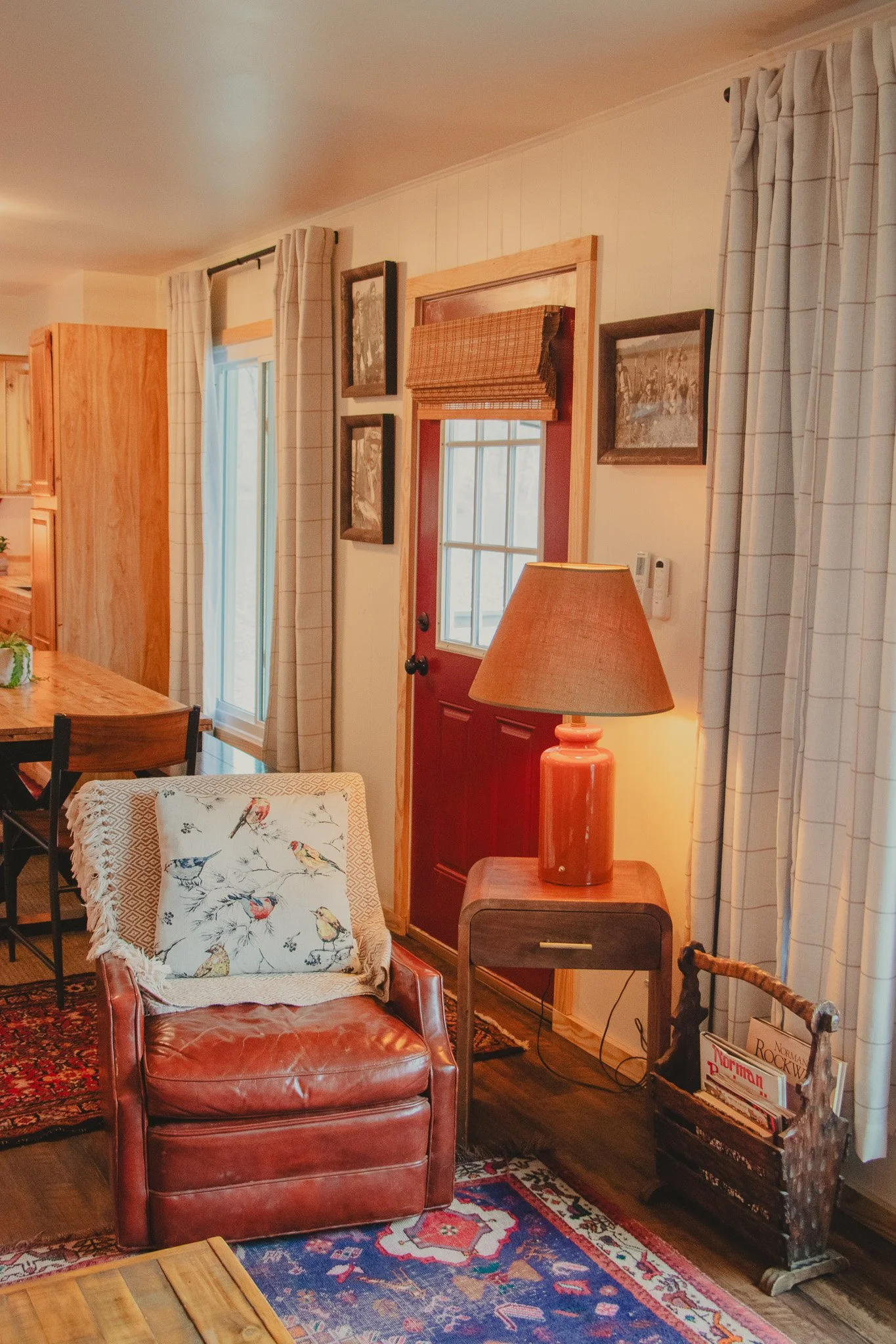 Living room corner with red door, beige curtains, table lamp, armchair with bird-patterned pillow, wooden side table, and magazine holder.