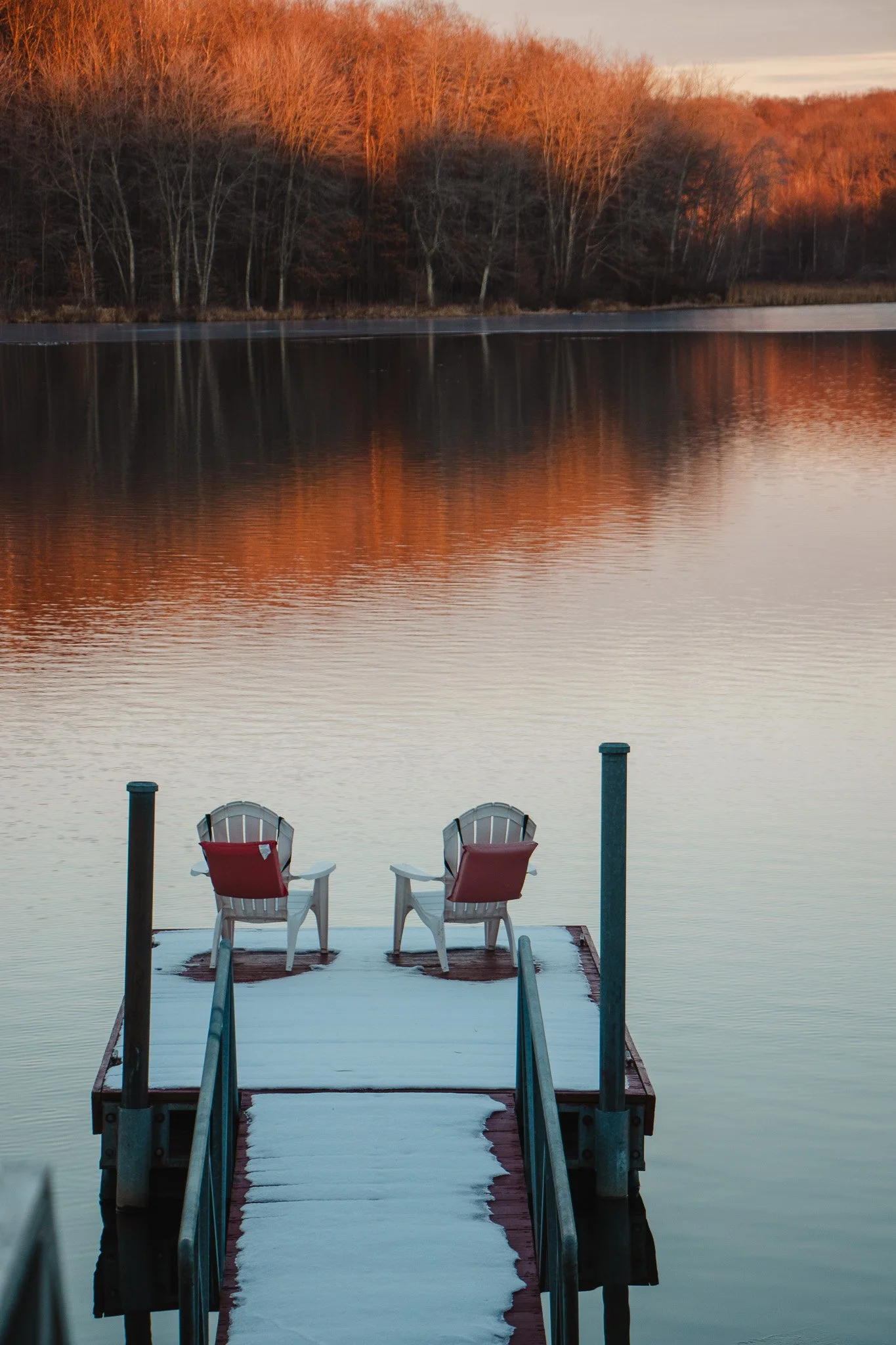 A snow-covered dock extends into a calm lake with two empty chairs facing the water, surrounded by leafless trees at sunset.