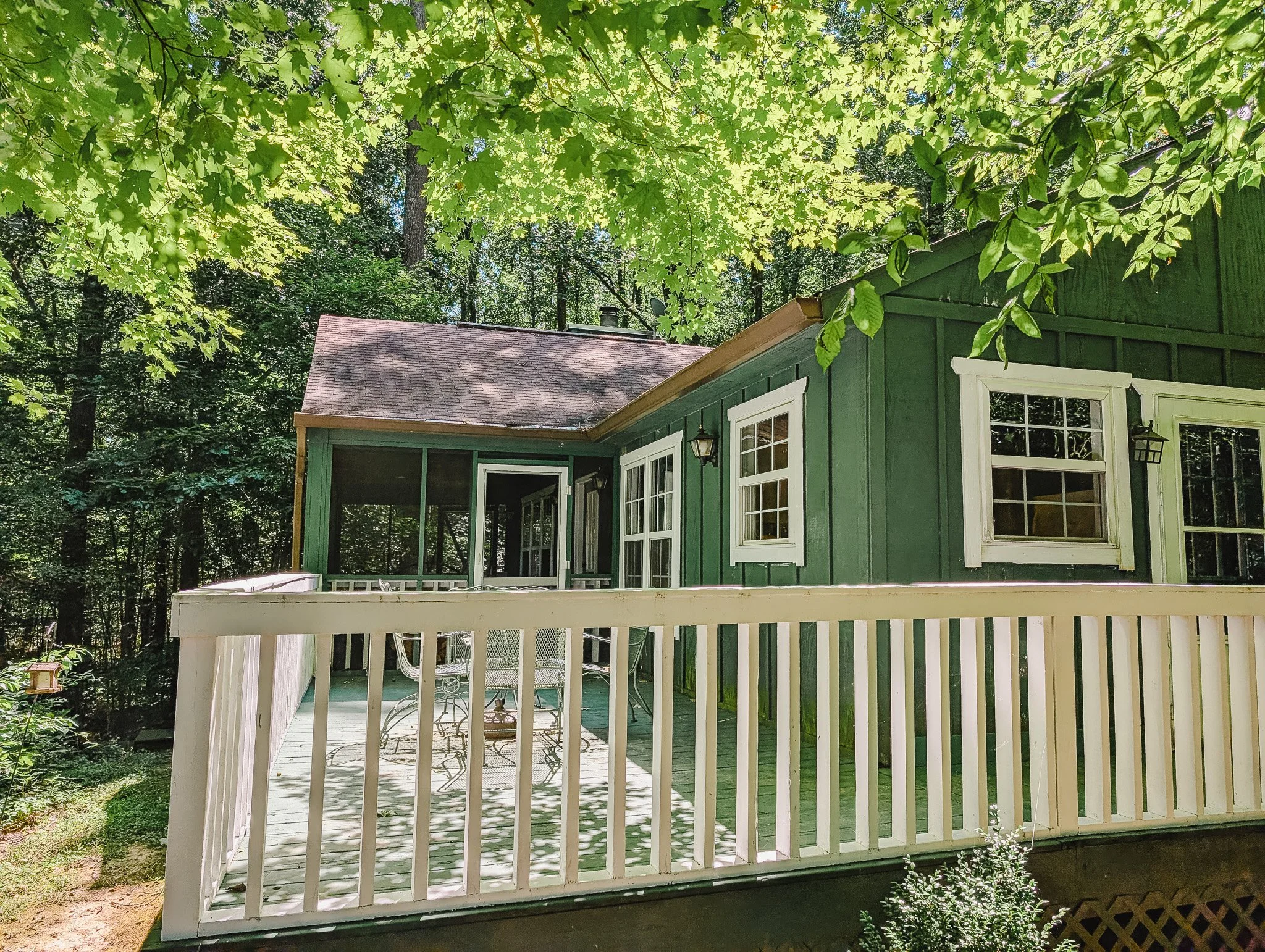 Green house with white window frames and a white railing, surrounded by tall trees with green leaves.