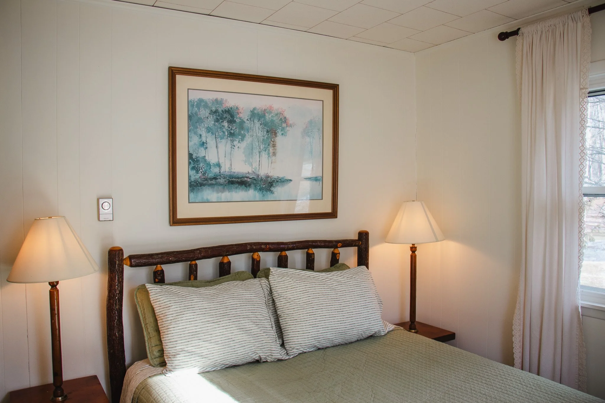 A cozy bedroom with a wooden bed frame, striped pillows, a green bedspread, two bedside lamps, a framed painting of trees, white walls, and a window with white curtains.