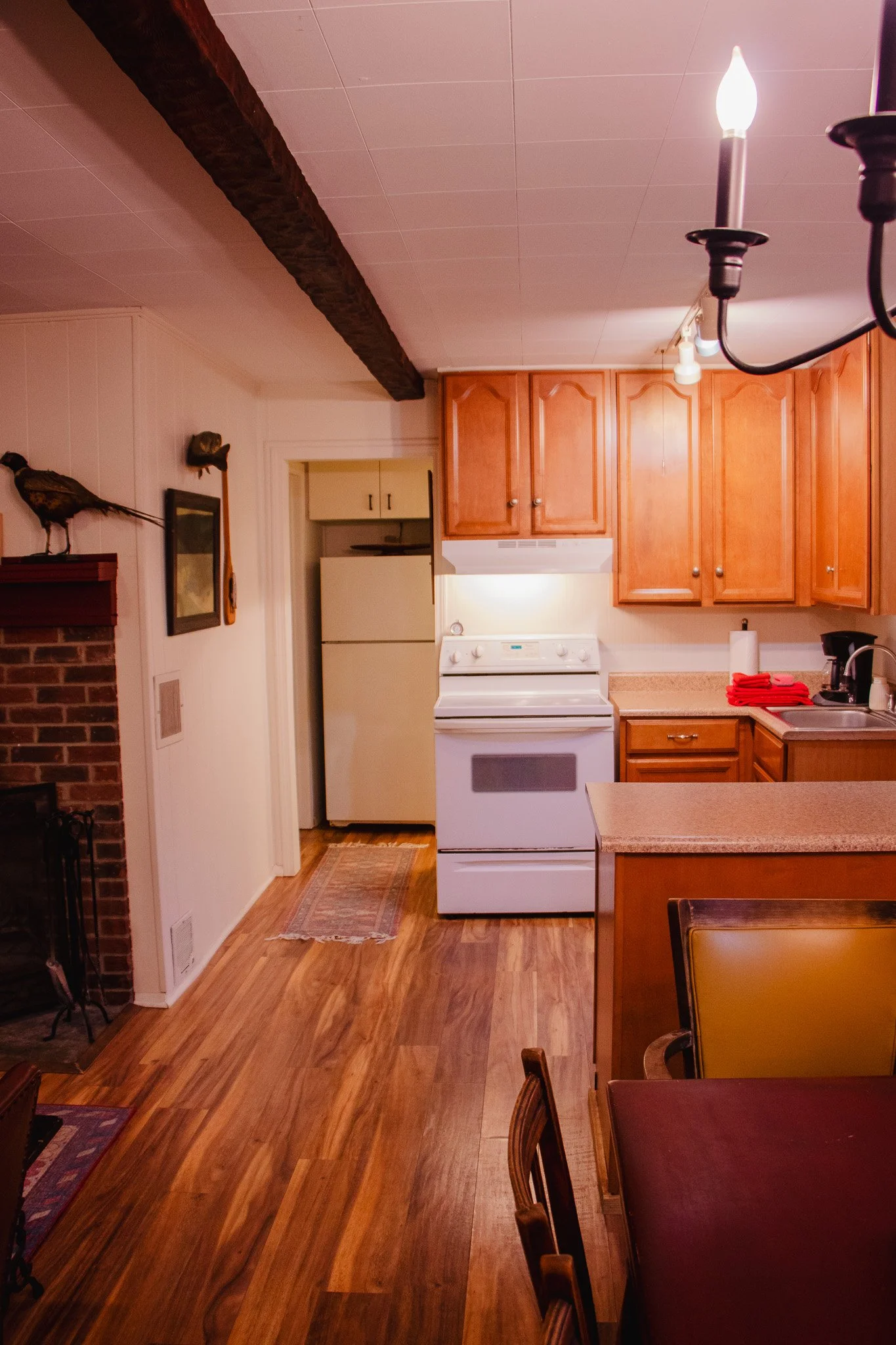 A cozy kitchen with wooden cabinets, white stove, refrigerator, a sink with red towels, and a dining table with chairs. There is a decorative bird sculpture on the brick fireplace, and wooden flooring throughout.