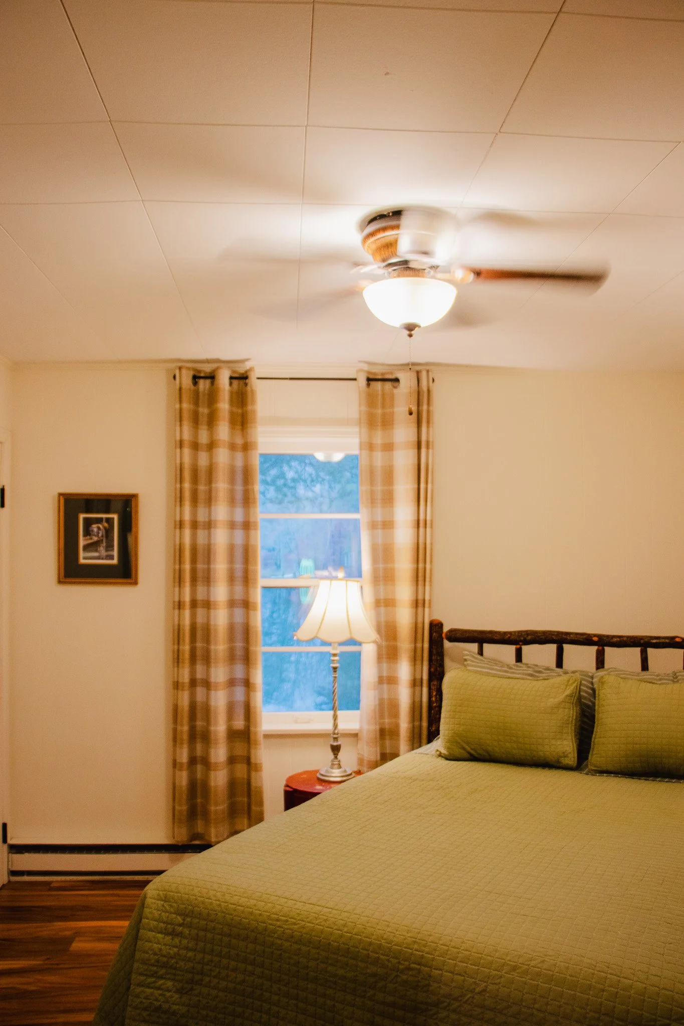 A cozy bedroom with a bed covered in a green quilt and matching pillows, a window with beige checkered curtains, a lamp on a small red table, a ceiling fan, and a framed picture on the wall.