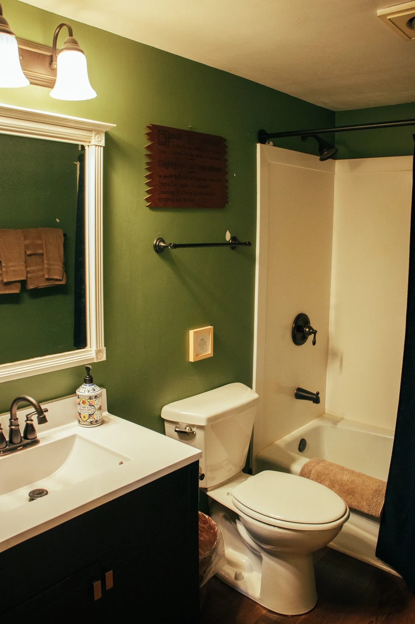 A small bathroom featuring a white sink with a dark cabinet, a mirror framed in white, a white toilet, and a bathtub with a dark shower curtain. The walls are painted green, with a towel rack holding three towels, a decorative soap dispenser, and som
