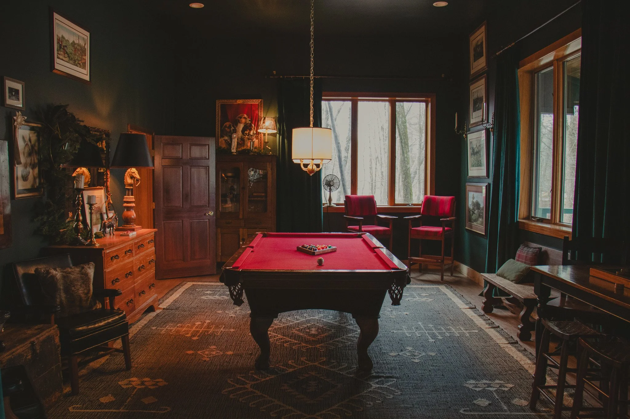 A cozy room with a red pool table in the center, surrounded by vintage furniture and artwork, with windows overlooking trees outside.