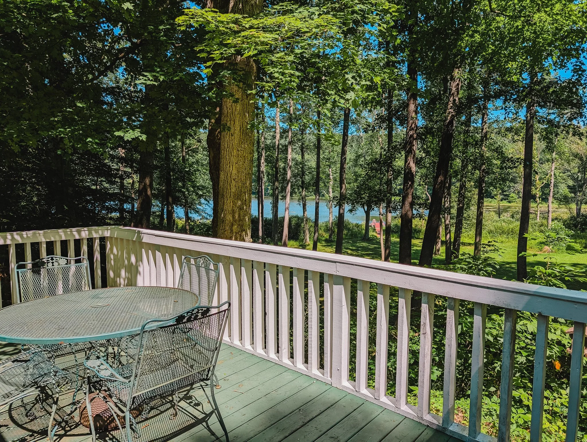 A wooden deck overlooking a wooded area with tall trees and a view of a lake in the background. There is a metal table and four chairs on the deck.