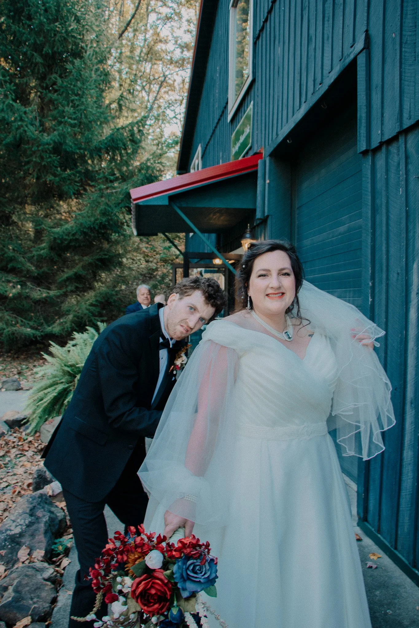 A bride in a white wedding dress holding a bouquet of colorful flowers, standing outdoors beside a groom in a black tuxedo, with a dark blue building and trees in the background.