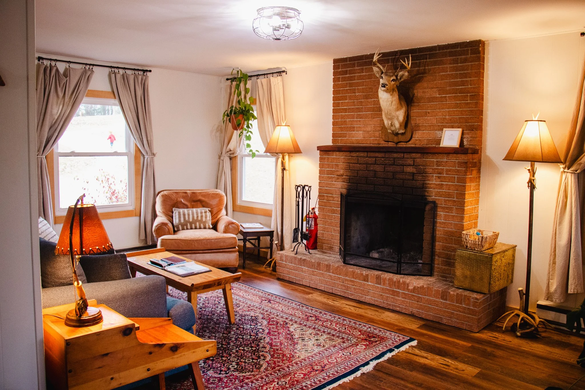 Living room with brick fireplace, mounted deer head, beige curtains, armchair, sofa, wooden coffee table, patterned rug, and standing lamps.
