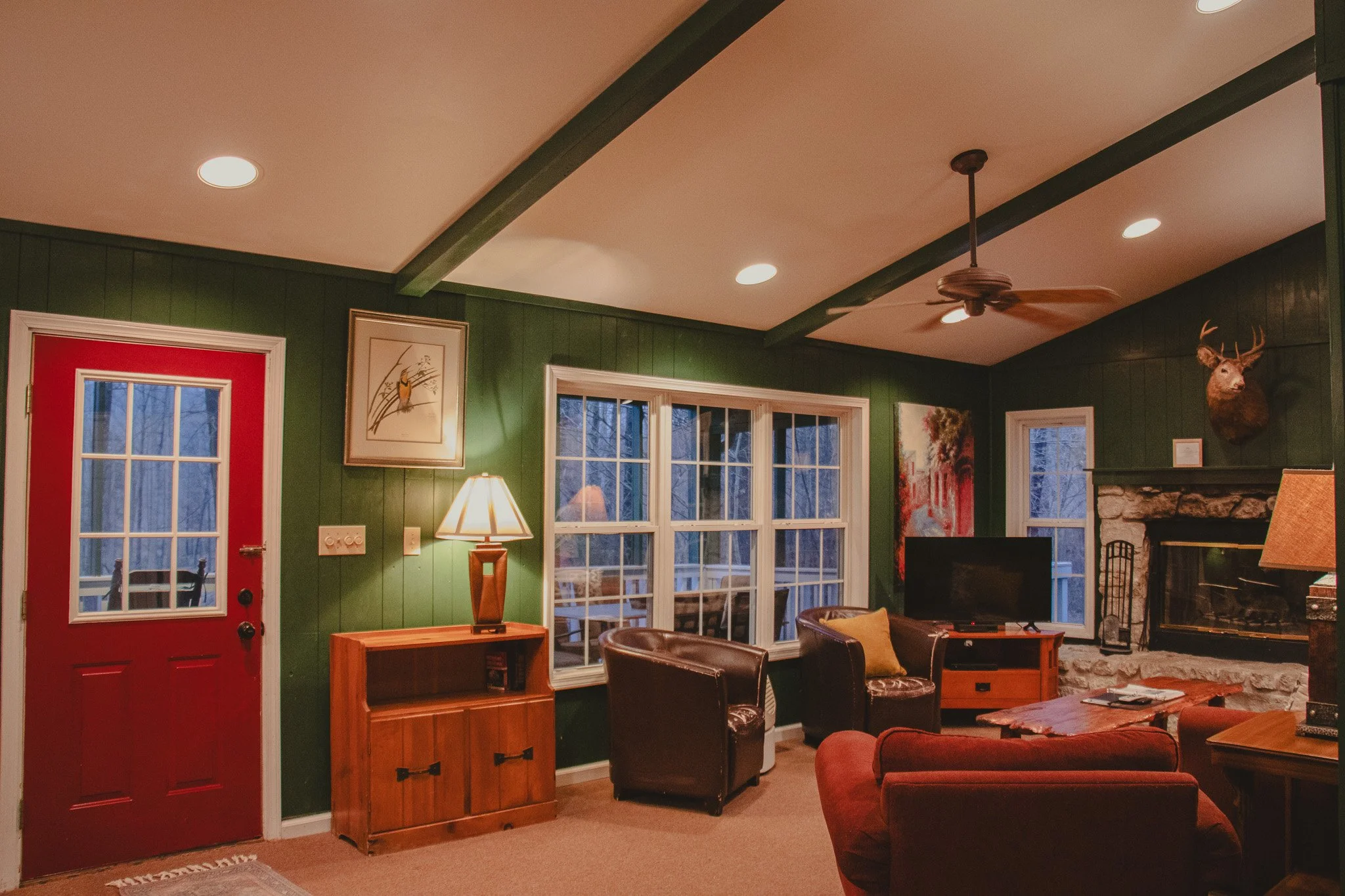 A cozy living room with dark green paneled walls, a red door with glass panes, a stone fireplace with a mounted deer head, and leather armchairs. There is a wooden TV stand and a small wooden side table with a lamp. Large windows behind the furniture