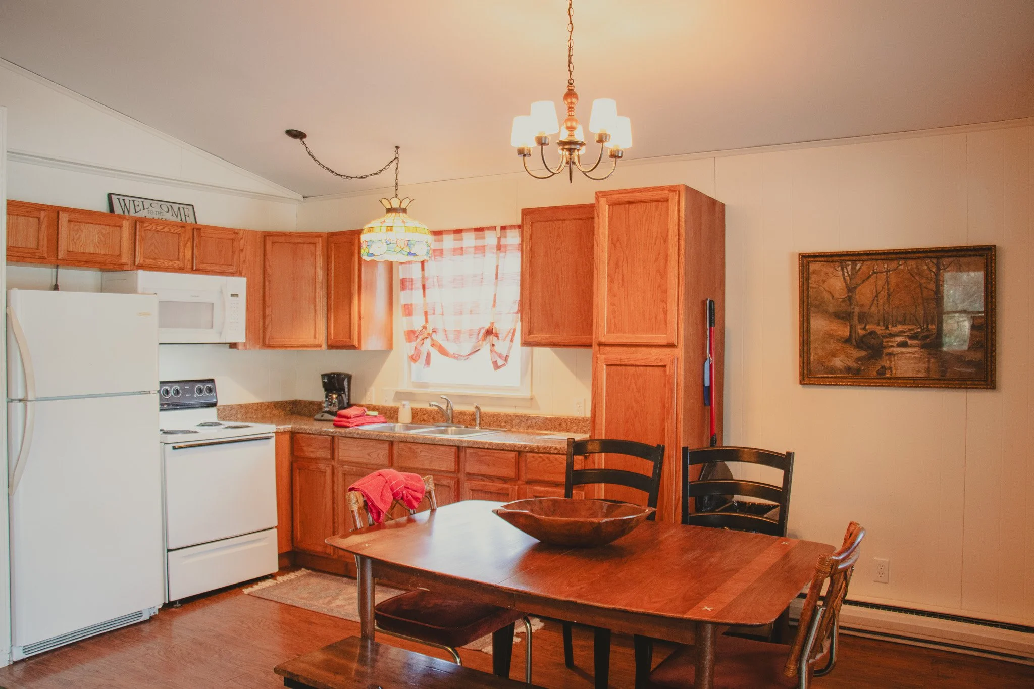 Kitchen with wooden cabinets, white appliances, and a wooden dining table with chairs, decor includes a wall painting and a 'Welcome' sign.