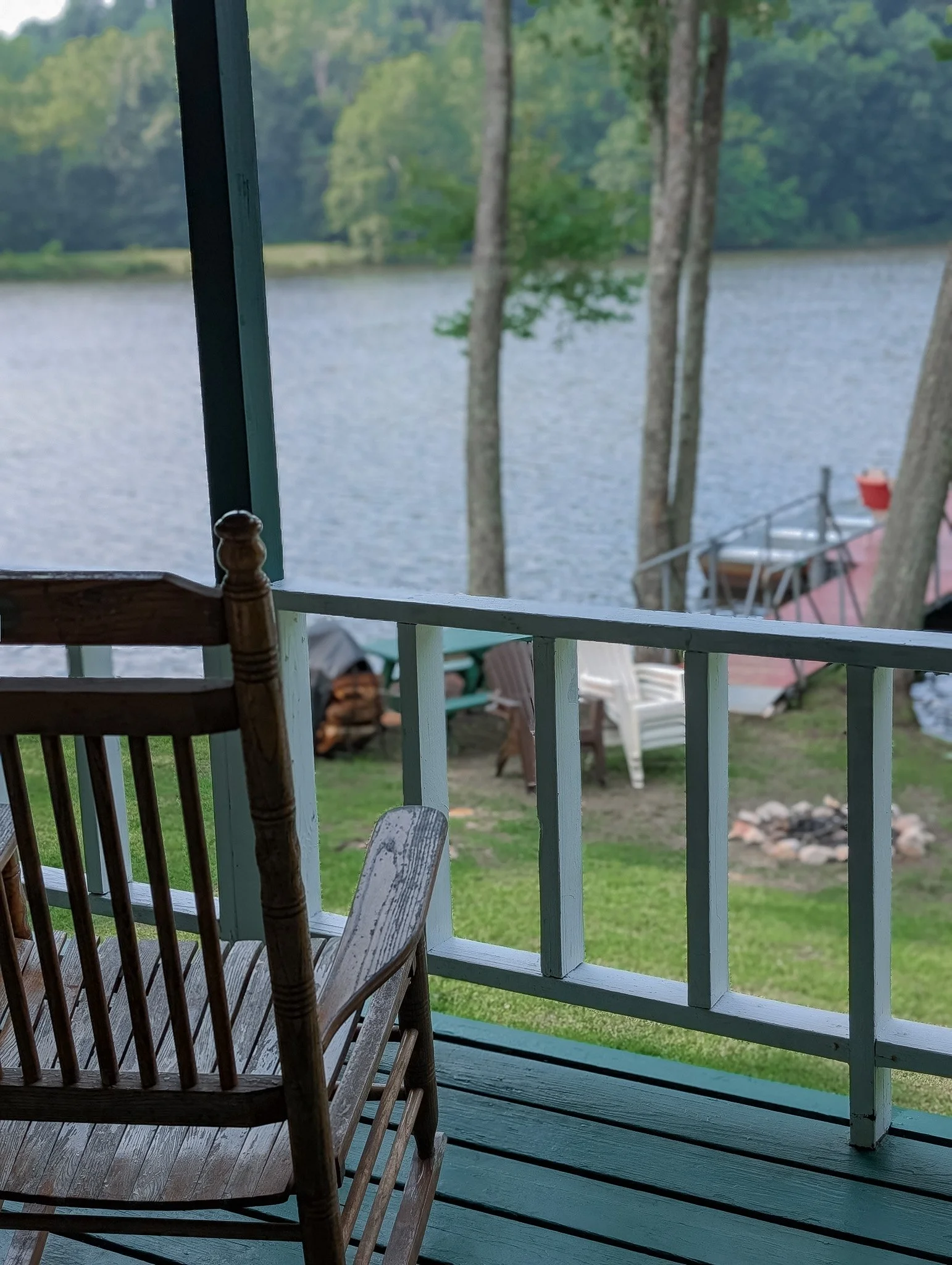 View from a porch overlooking a lake with trees, chairs, and a fire pit in the yard.