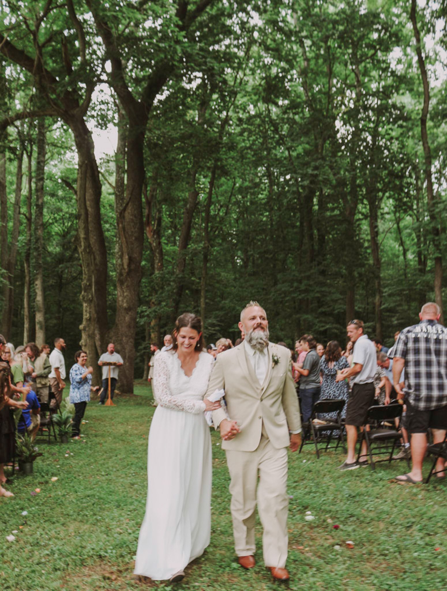A bride and groom walking down the aisle during their outdoor wedding ceremony in a wooded area, smiling and holding hands.