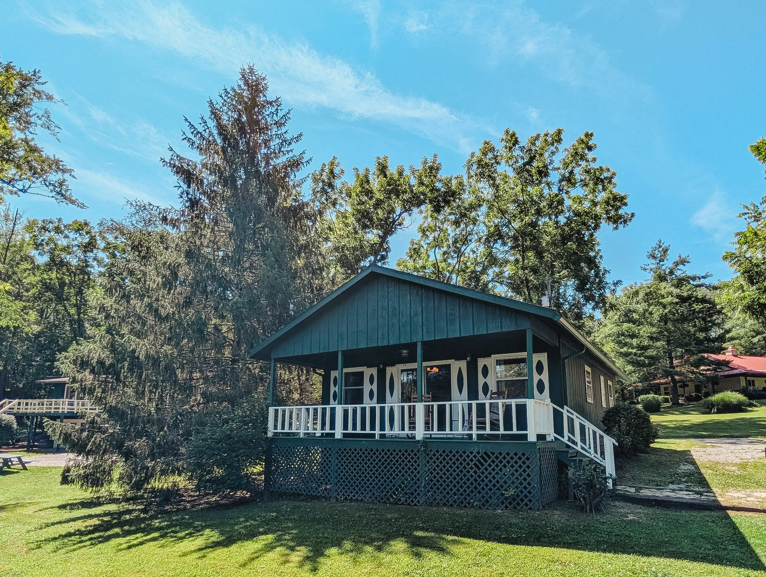 A house with a porch and stairs, surrounded by green grass and tall trees under a clear blue sky.