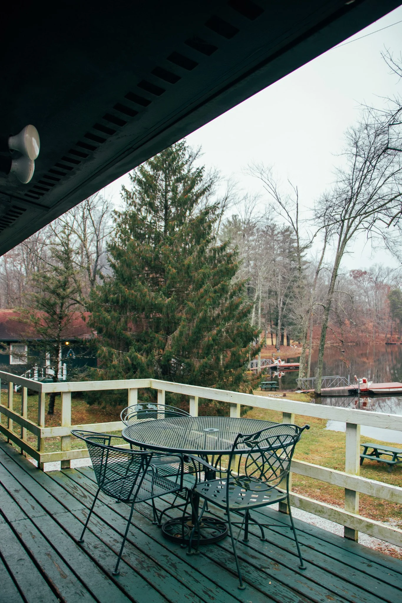 View from a covered porch with black metal outdoor furniture, overlooking a yard with a wooden railing, trees, and a lake with docks in the distance.