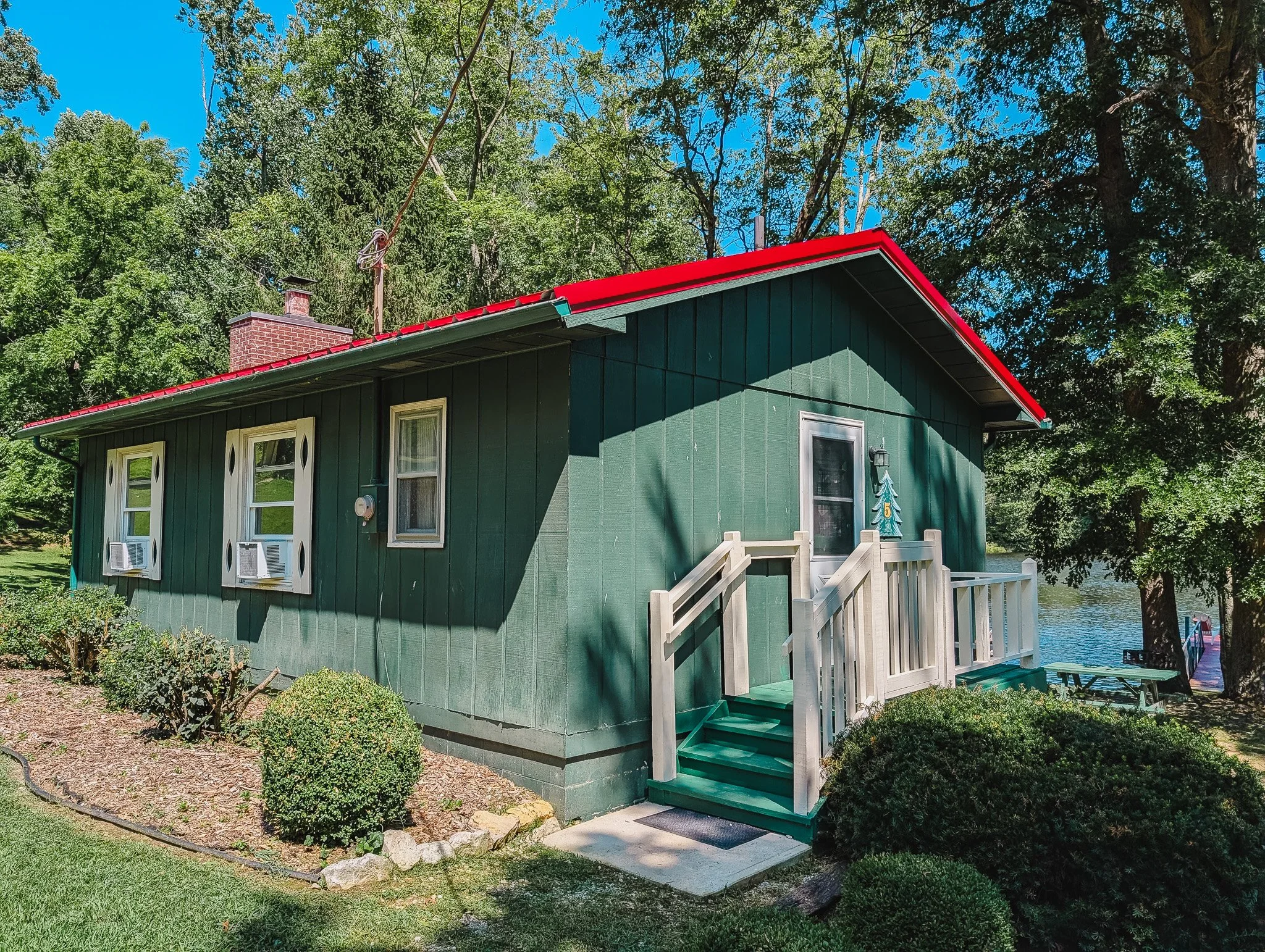 A small green house with a red roof located near a body of water, surrounded by trees and bushes.