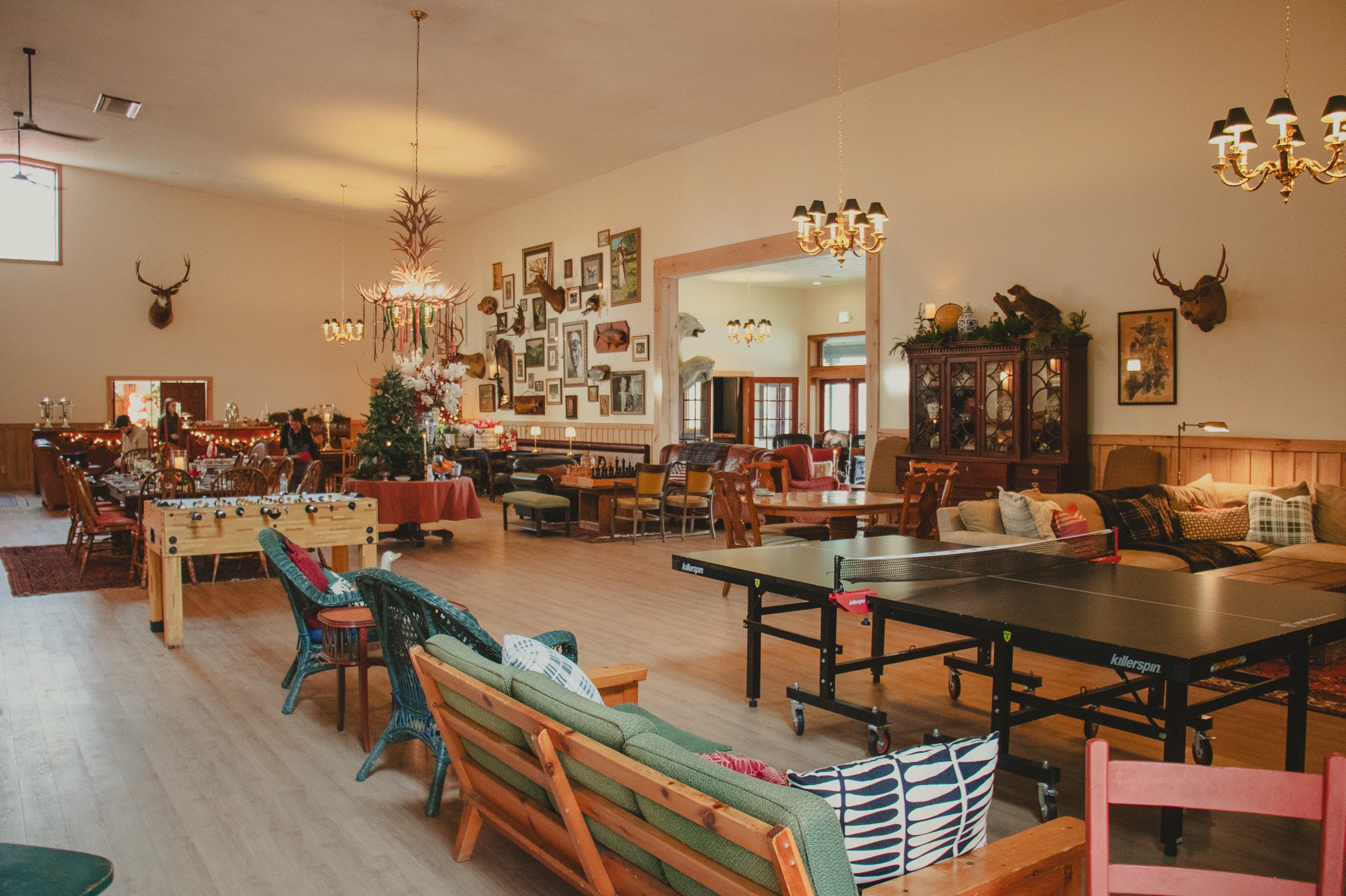 A spacious room decorated with ecological and rustic theme, featuring animal mounts on the walls, framed pictures, and vintage furniture, with a ping pong table in the foreground and a Christmas tree in the center.