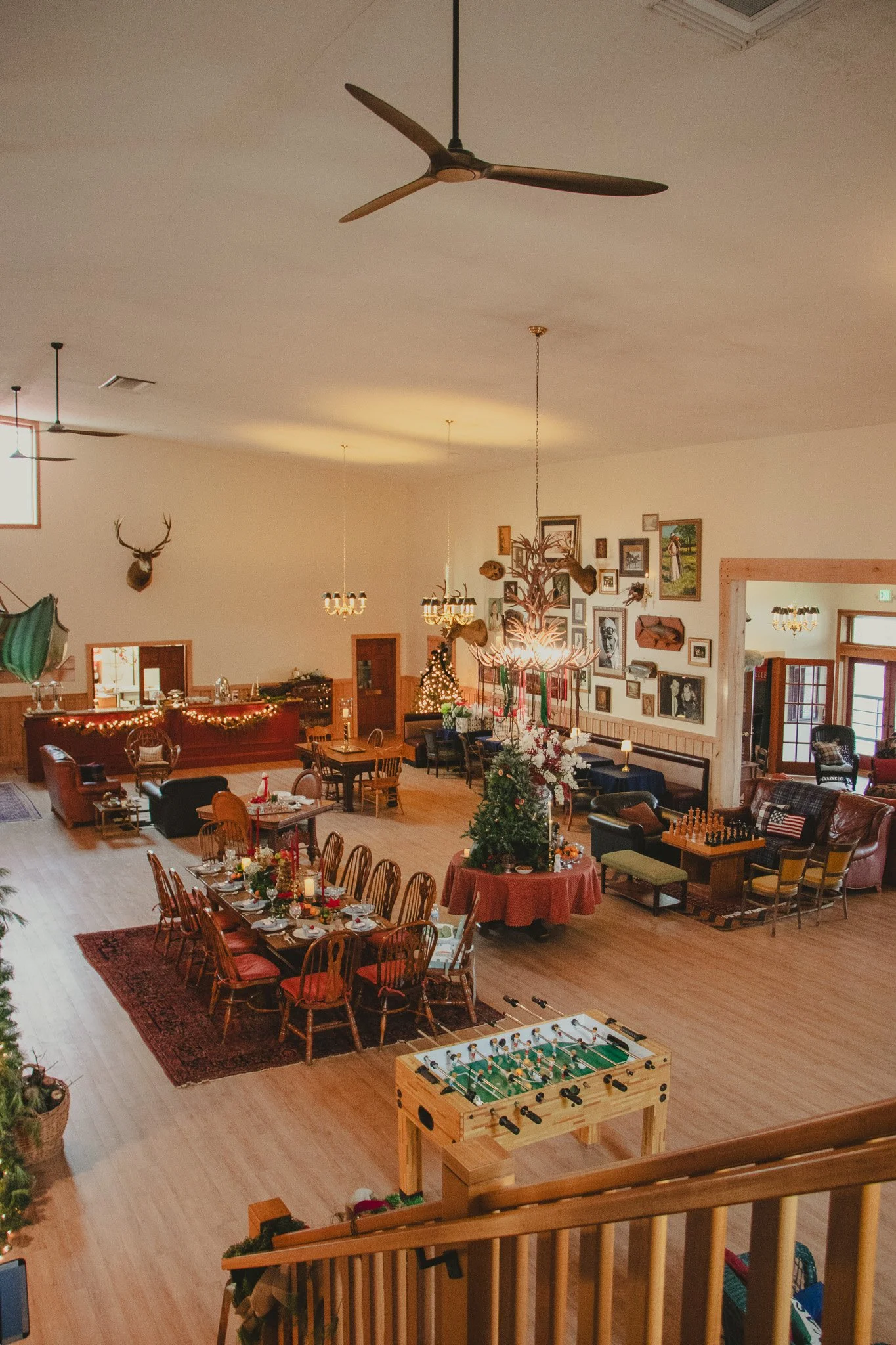 A spacious indoor room decorated for Christmas with a large dining table, a Christmas tree, and lounge areas featuring armchairs and a foosball table, with wall art and mounted animal heads.
