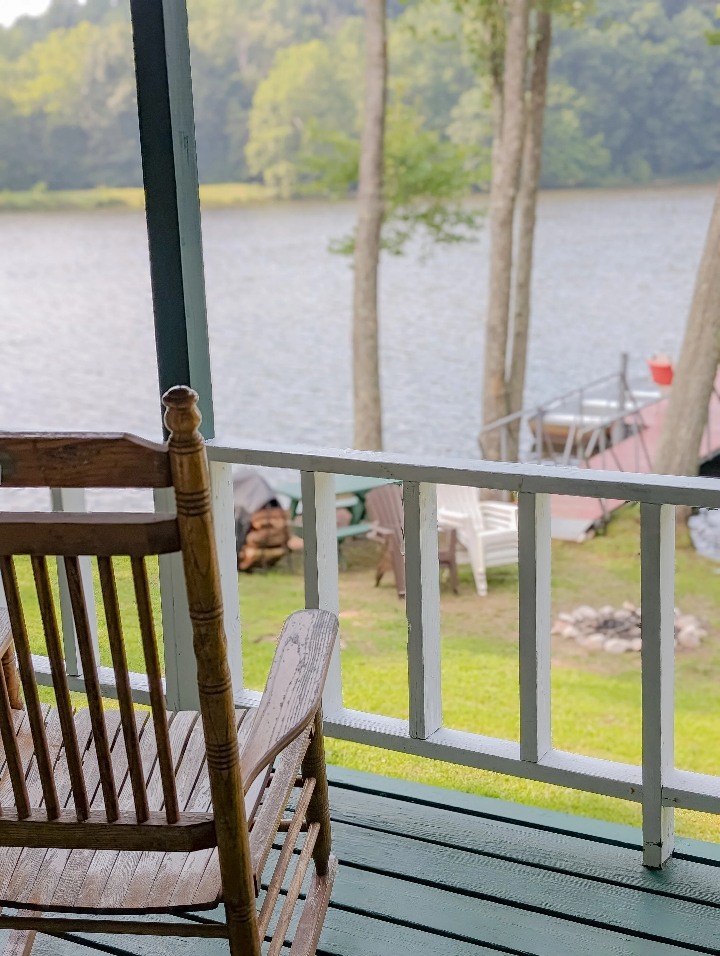 View of a peaceful lakeside scene from a porch, with a wooden chair in the foreground, trees and a dock extending into the lake in the background.