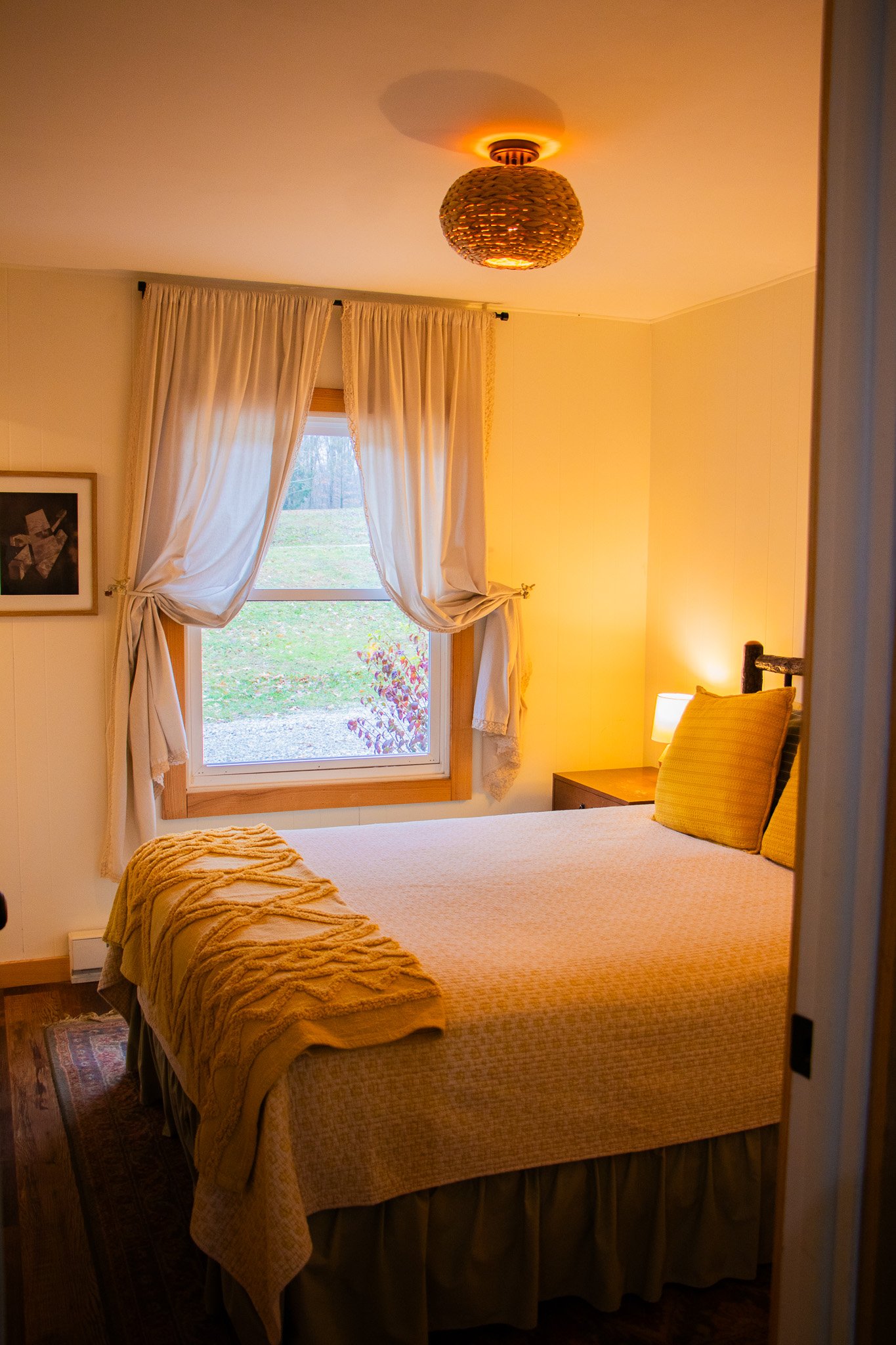 A cozy bedroom with a bed, yellow pillows, a blanket, a bedside lamp, a window with cream-colored curtains, and a view of greenery outside.