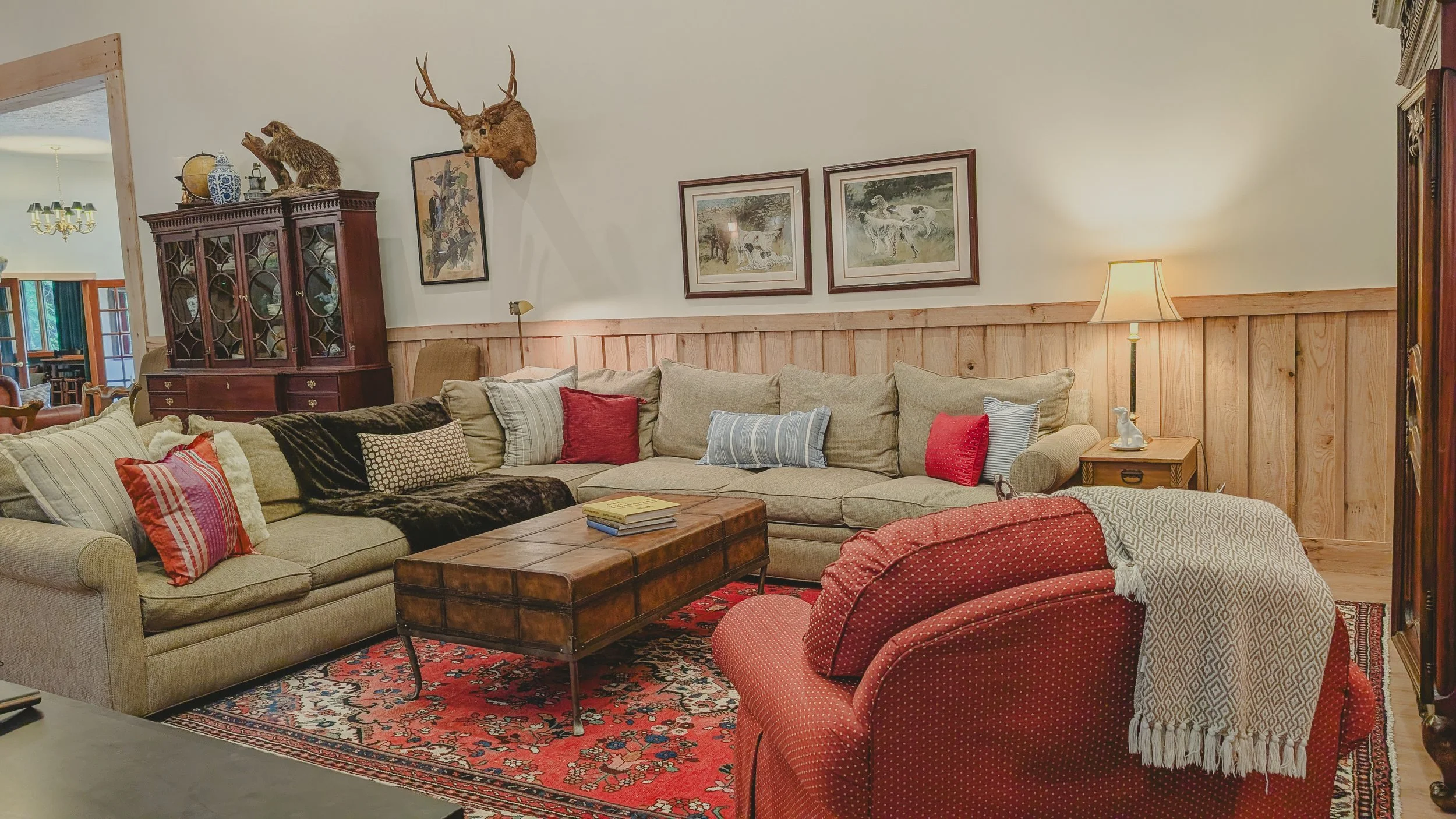 Living room with beige sectional sofa, red armchair, wooden coffee table, patterned area rug, wall art, wooden wall paneling, and decorative items including animal-themed mounted head and bear, and a cabinet displayed in background.