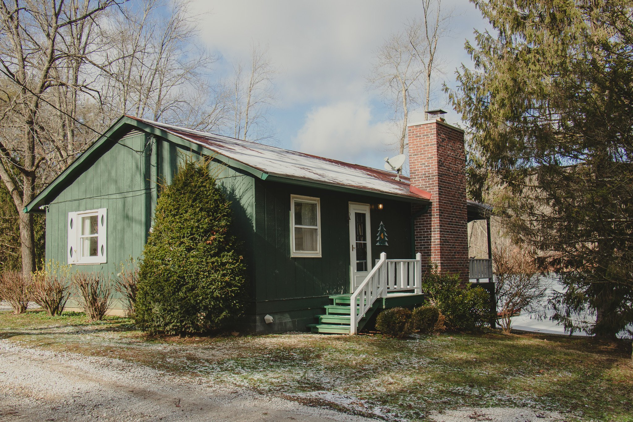 A small green house with white trim, a brick chimney, a porch with green stairs, and a Christmas tree decoration on the door, surrounded by trees during winter with some snow on the ground.