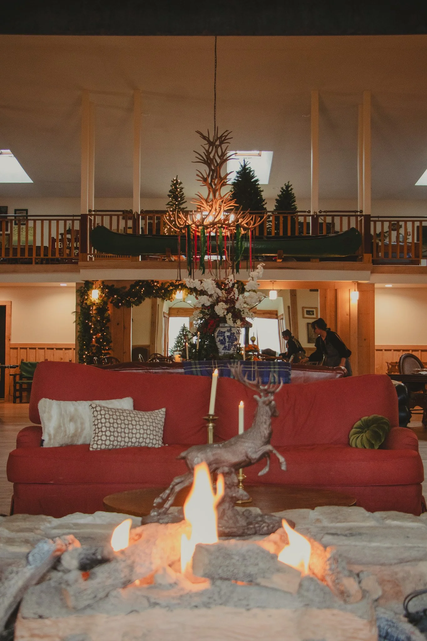 Cozy living room decorated for Christmas with a fire in the foreground, a red sofa with pillows, a large floral arrangement, and a chandelier made of antlers hanging from the ceiling.