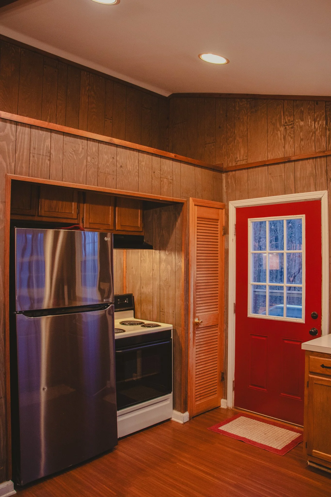 A kitchen corner with a red door, wooden walls, a refrigerator, a stove, and a small window on the door.