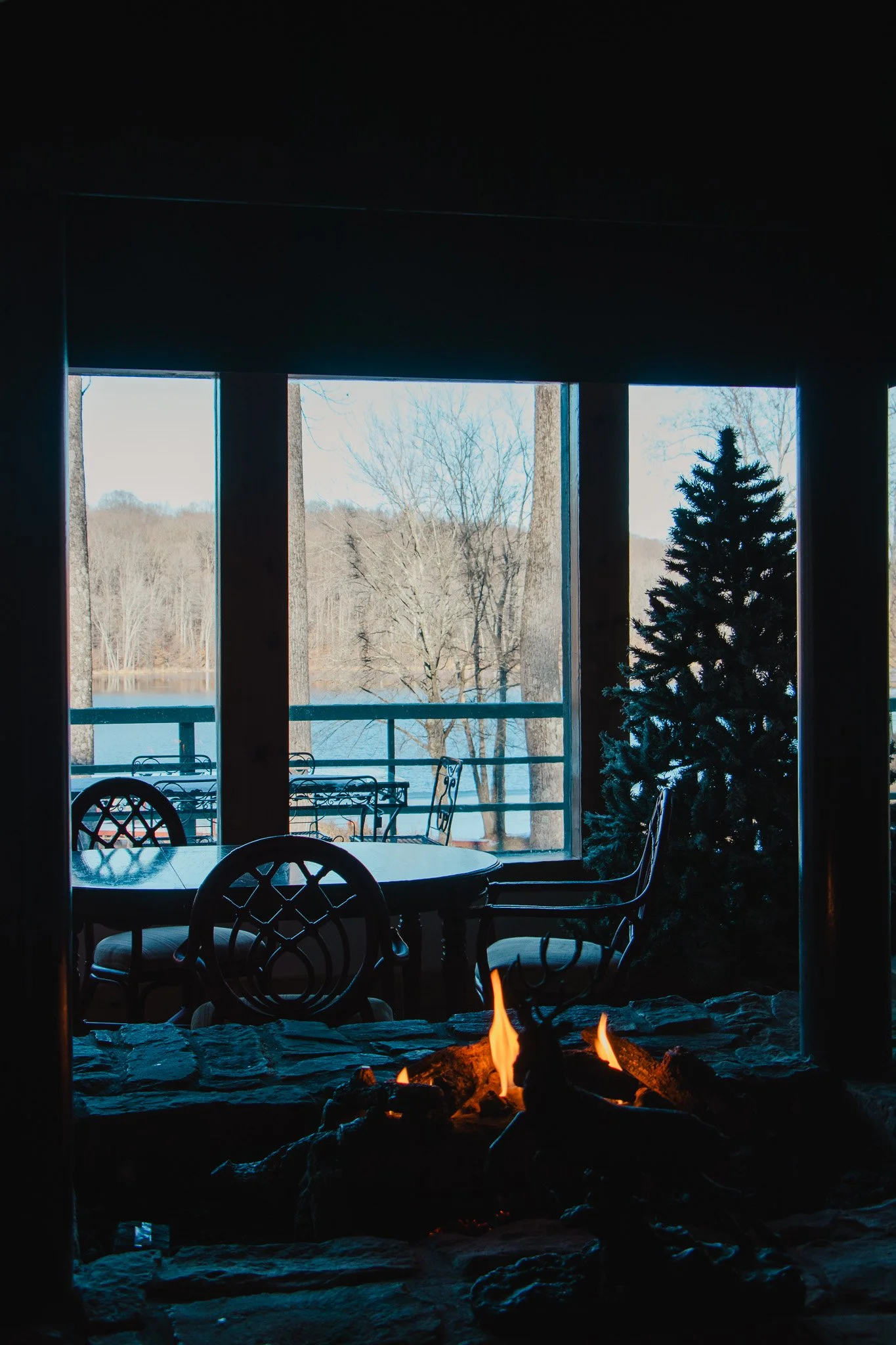 A cozy room with a fireplace, a Christmas tree, a wooden table, and chairs, overlooking a snowy outdoor landscape through large windows.