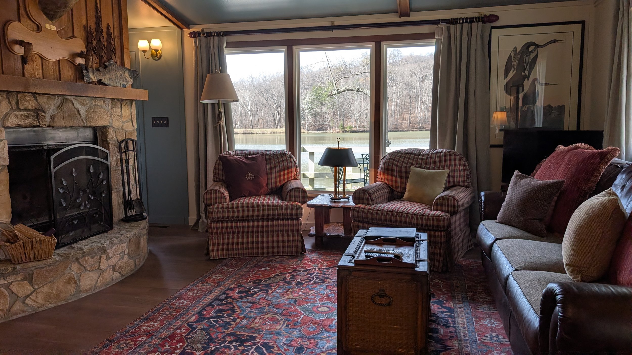 Cozy living room with a stone fireplace, mounted deer head, and wood-paneled wall. A leather armchair with an ottoman is near the fireplace, with a patterned rug on the wooden floor.