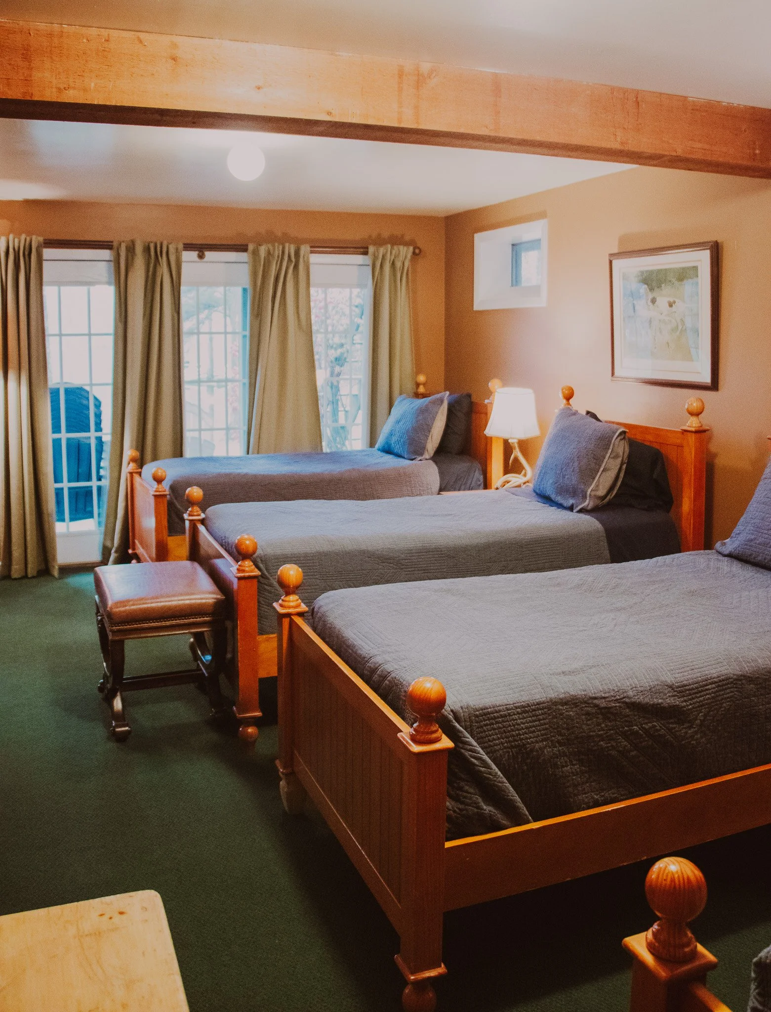 A cozy bedroom with three wooden beds with gray bedding, a lamp between two beds, a framed picture on the wall, and a glass sliding door with beige curtains leading outside.