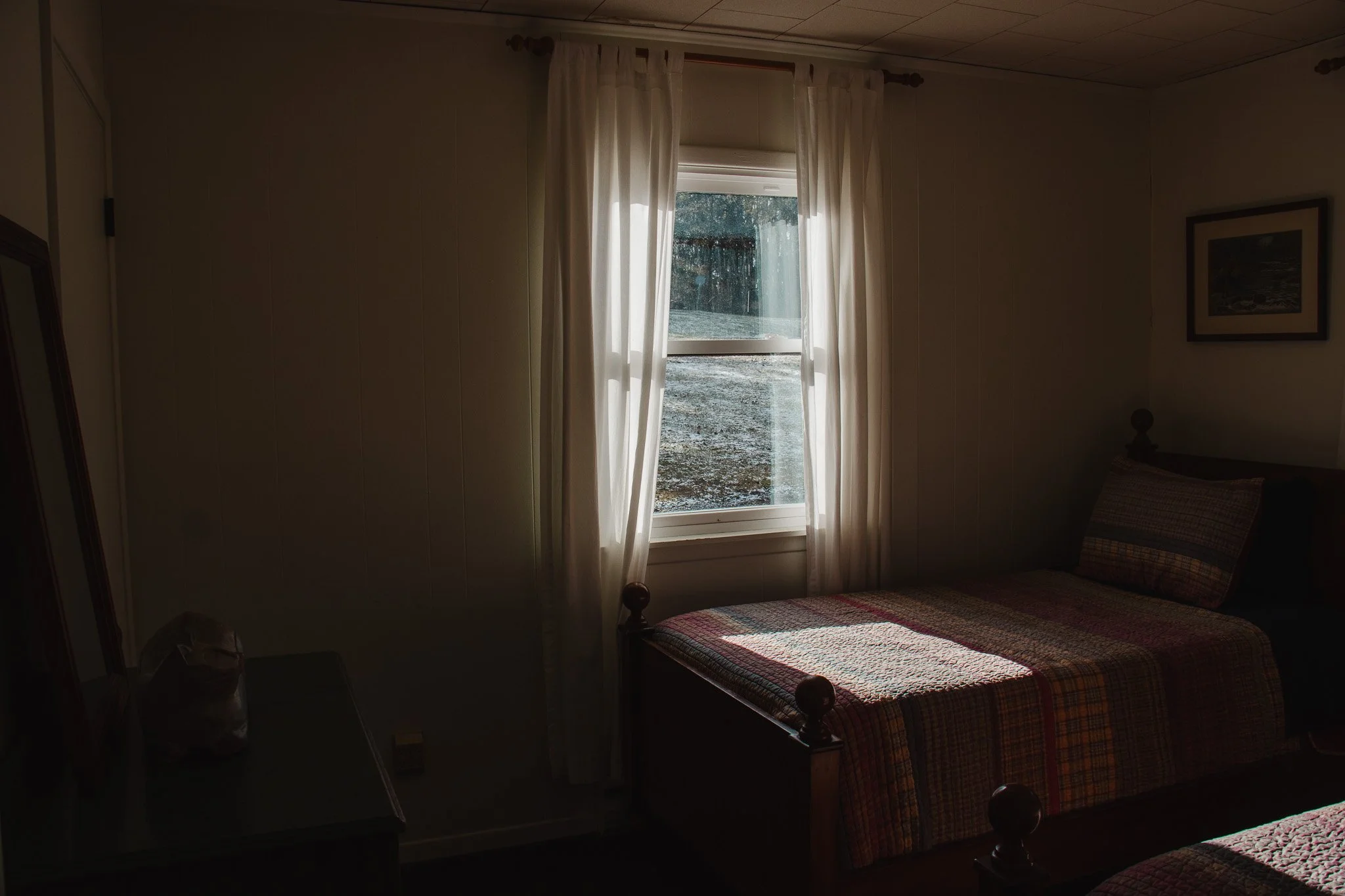 A bedroom featuring a window with sheer white curtains, a bed with a colorful checkered quilt, and a framed picture on the wall.