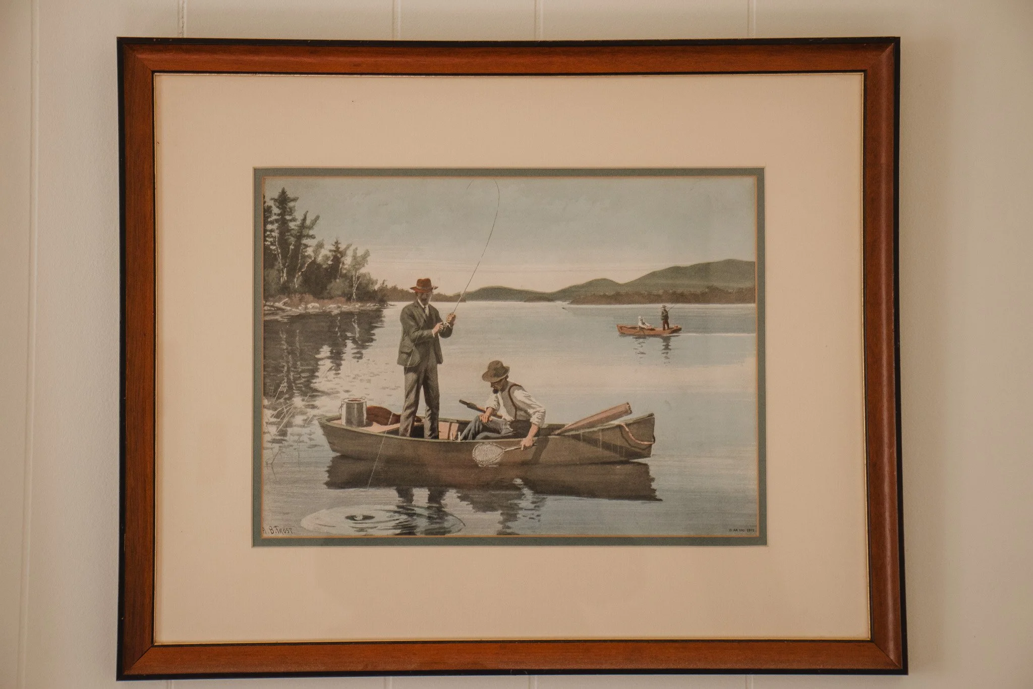 A framed painting of two men fishing in a boat, with a calm lake, trees, hills, and a smaller boat with two people in the background.