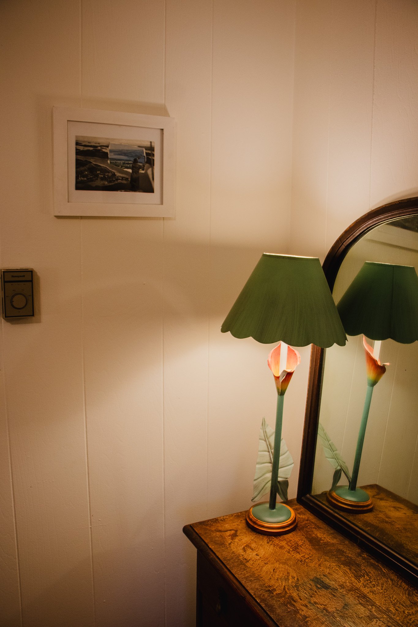 A wooden dresser with a mirror and a floral table lamp with a scalloped green shade. A small framed landscape photo hangs on the beige wall above.