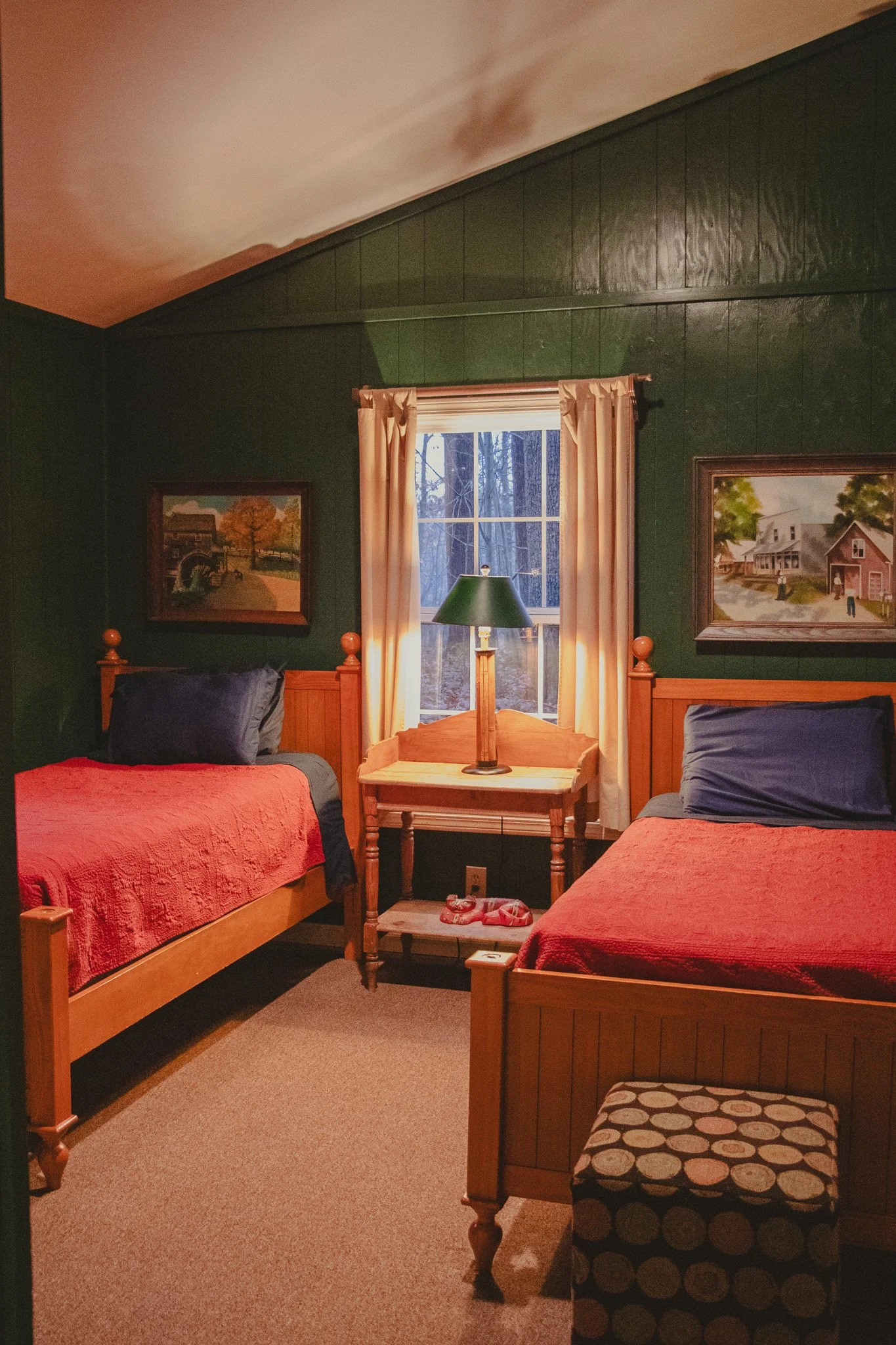 A cozy bedroom with dark green wooden walls, two wooden twin beds with red quilts and navy pillows, a small wooden table with a green-lamp between the beds, a window with cream-colored curtains, and framed paintings on the wall.