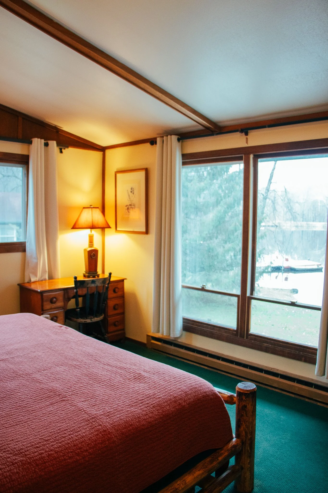 A cozy bedroom with a bed covered in a pink quilt, a wooden desk with a lamp and an office chair, beige curtains, and large windows showing a snowy outdoor scene.