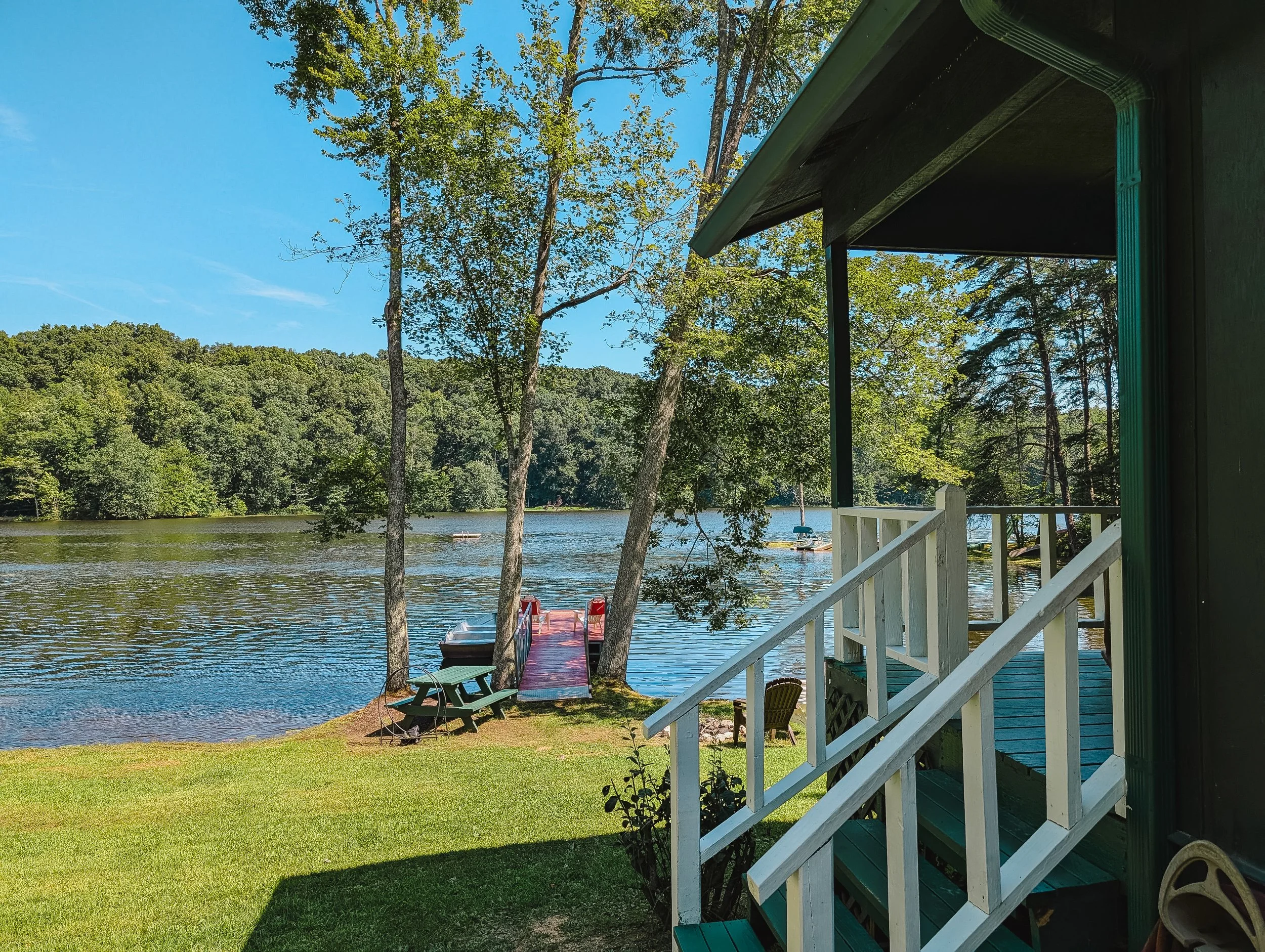 View from a porch overlooking a lake with a red dock, boats, and a lush green forest in the background on a sunny day.