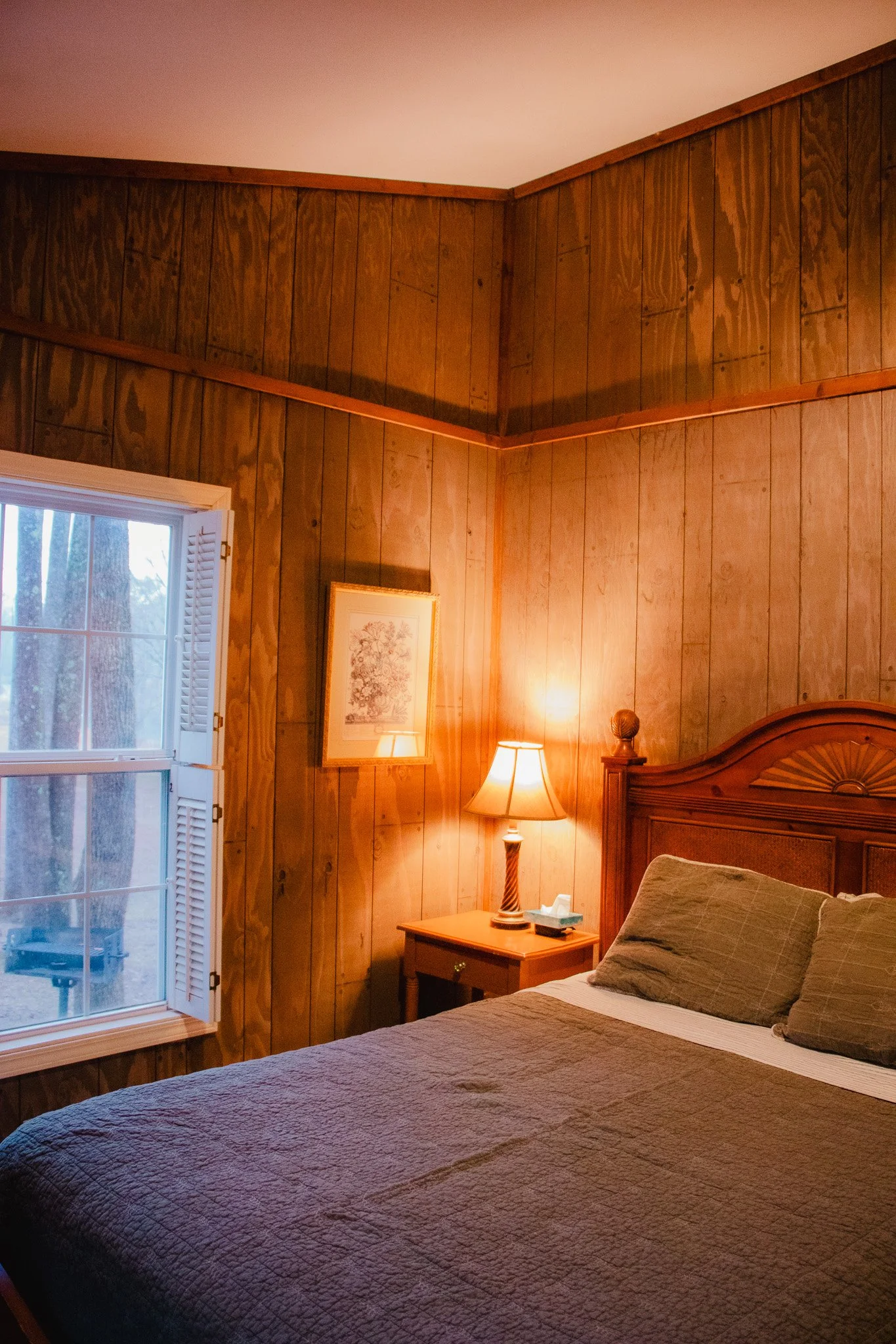 A cozy bedroom with wood-paneled walls, a window with white shutters, a wooden headboard, and a nightstand with a lamp.