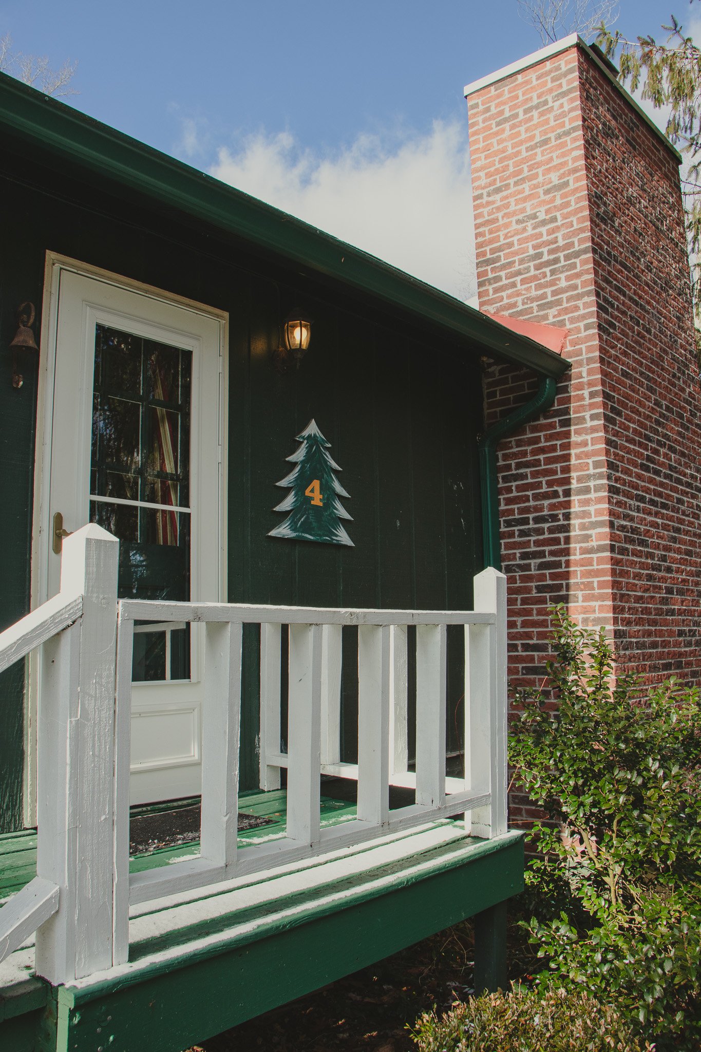 Front porch of a house with a white door, a dark exterior wall, a painted Christmas tree with the number 4 on it, and a red brick chimney, under a partly cloudy sky.