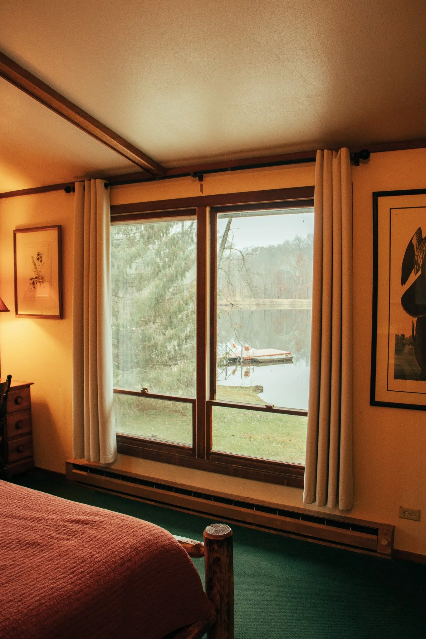 A bedroom window with white curtains showing a view of a lake, boat, and trees outside.