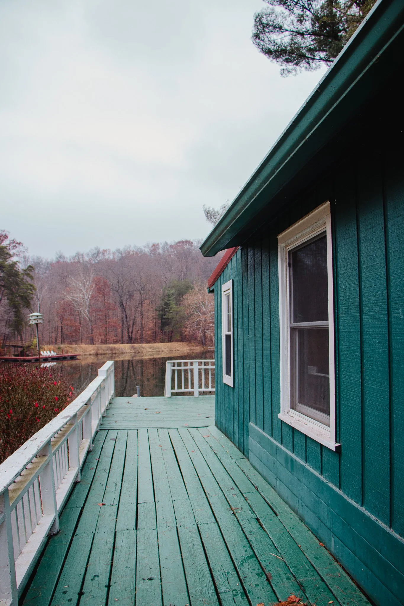 A wooden deck attached to a teal house with white trim, overlooking a lake with trees in the background, under an overcast sky.