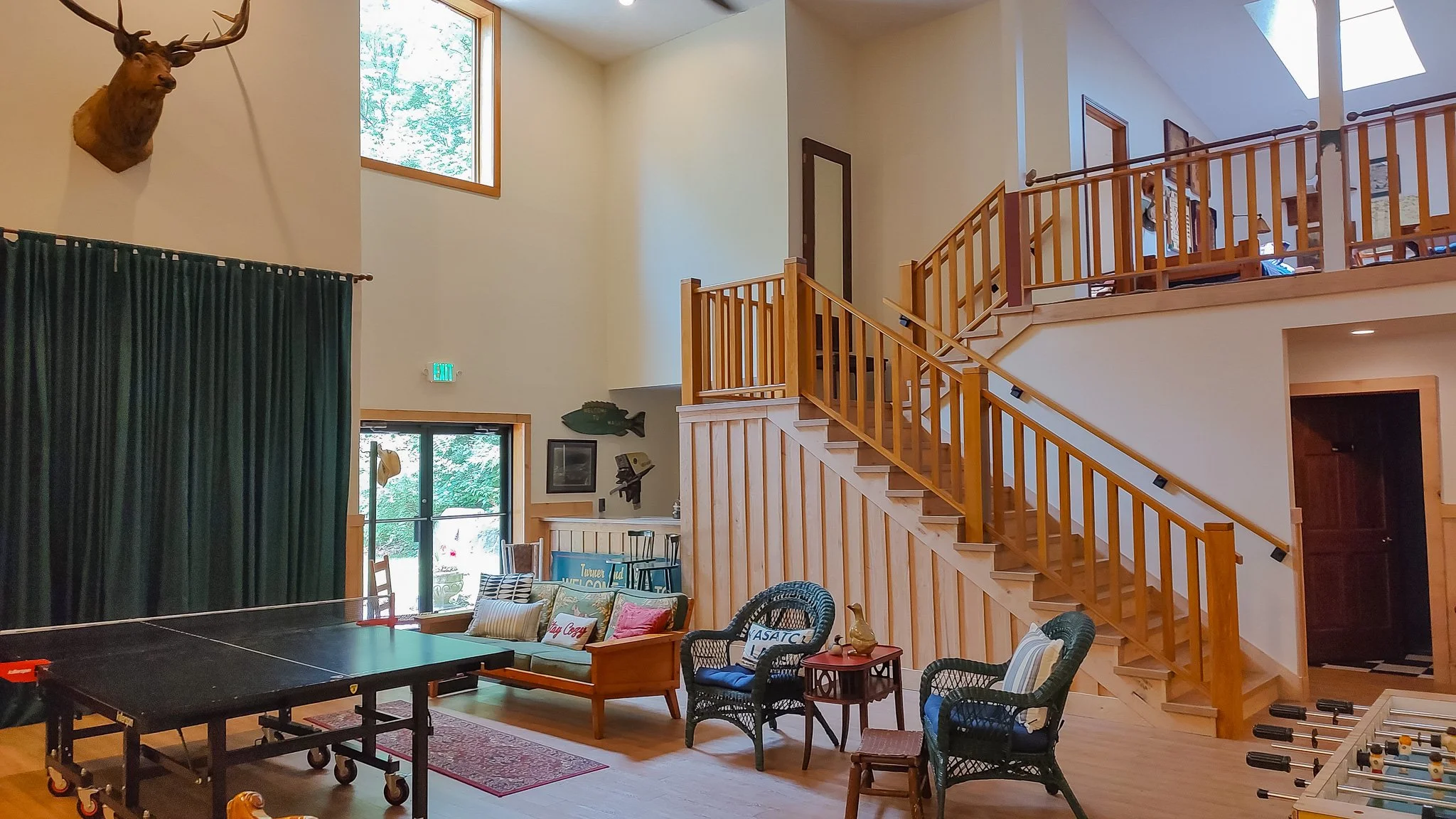 Interior of a spacious, two-story room with wood paneling, a staircase with wooden railing, a mounted deer head on the wall, a large window, and various seating and recreational furniture.