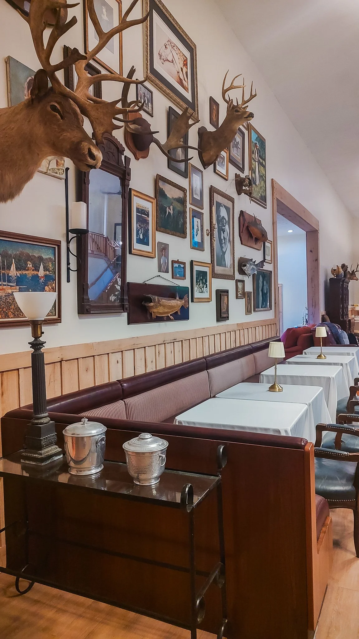 Interior of a restaurant with a collage of framed pictures and mounted deer and fish heads on the wall, wood-paneled section, tables with white tablecloths and small lamps, and a wooden sideboard with silver containers.