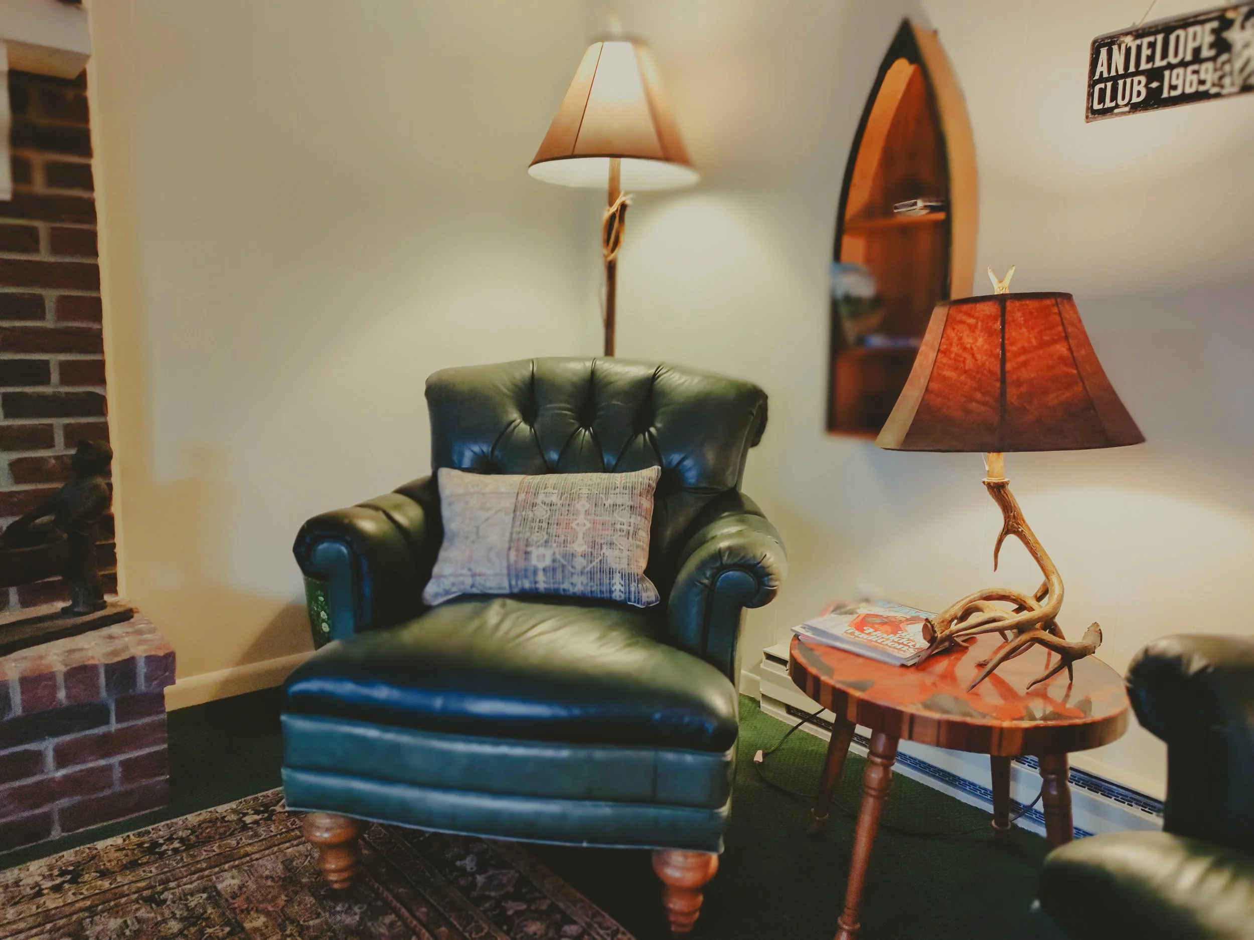 Living room with a green leather armchair, a wooden side table with a lamp and magazines, a wall mirror, and a brick fireplace with a small statue.