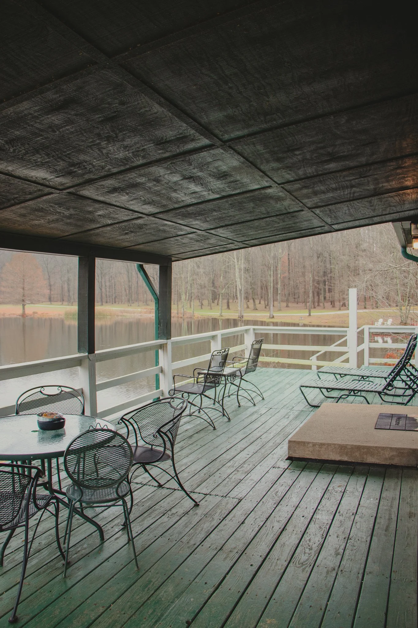 Empty outdoor patio with metal tables and chairs overlooking a lake, with a wooded area in the background.