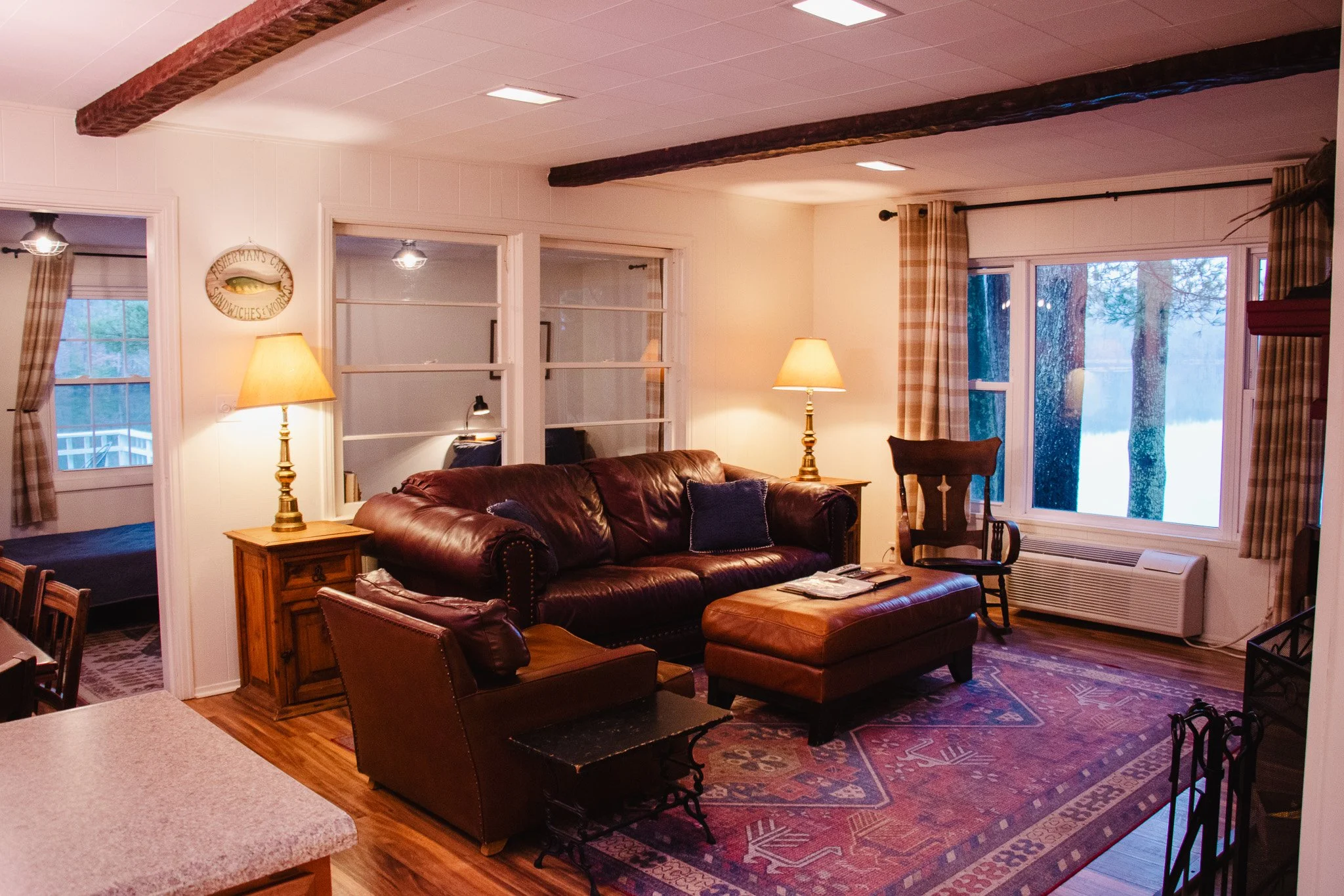 Cozy living room with leather sofa, wooden side tables with lamps, a rocking chair, large windows with checkered curtains, and an area rug on hardwood floor.