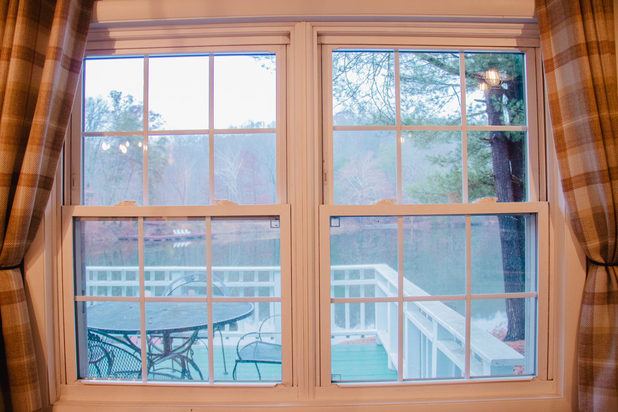 View through a large indoor window showing an outdoor patio with a round table and three chairs, surrounded by trees and a railing, with a lake or pond in the background.