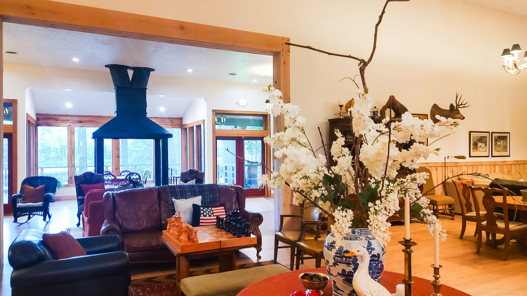 Living room with wooden accents, large windows, and a fireplace. Foreground features a table with a large floral arrangement, a swan-shaped ornament, and two tall candles. Furniture includes leather chairs and a sofa with pillows, and a chess set on 